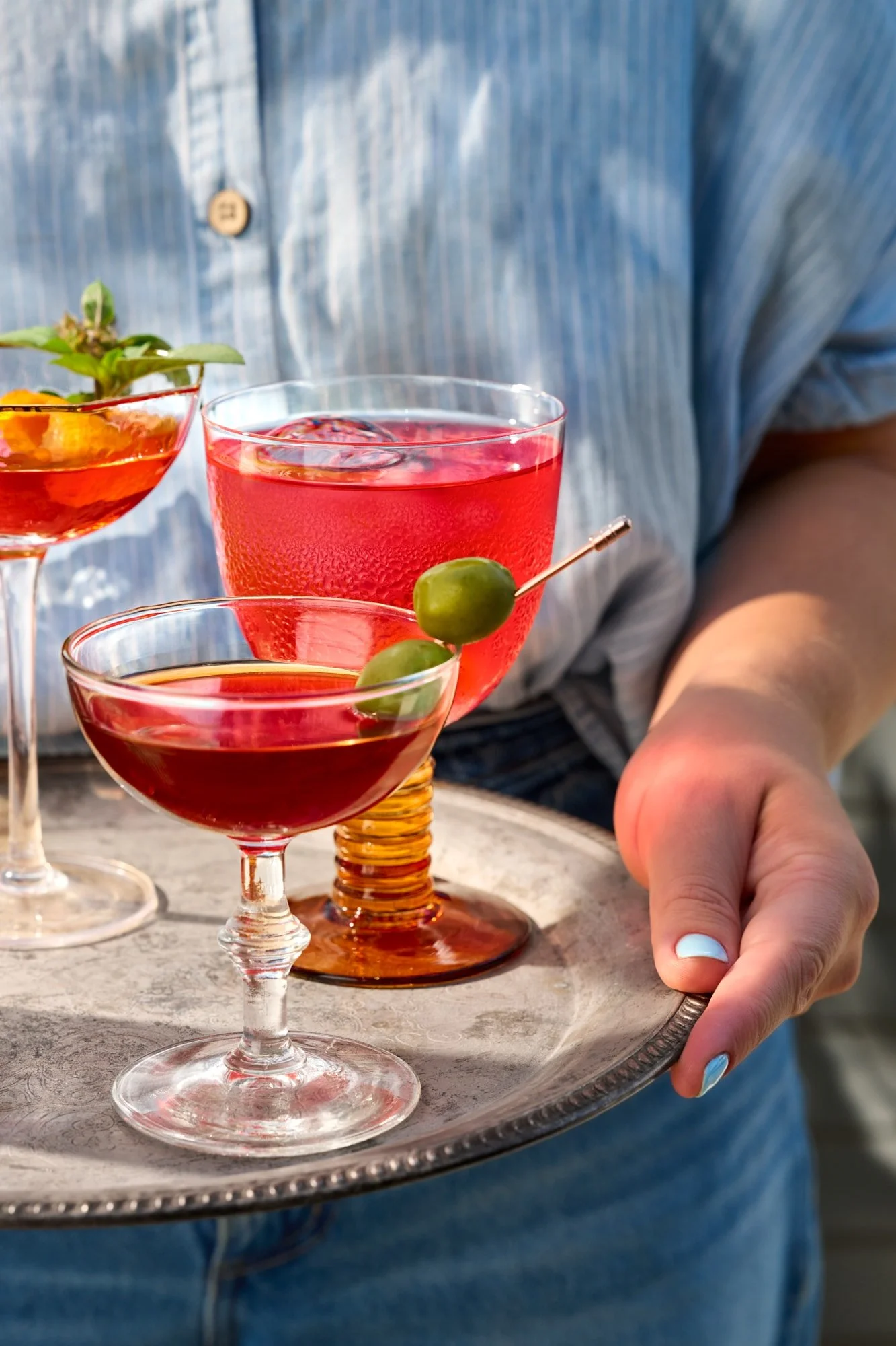Three colorful cocktails on a serving tray, including a red drink garnished with green olives and an orange cocktail with herbs.