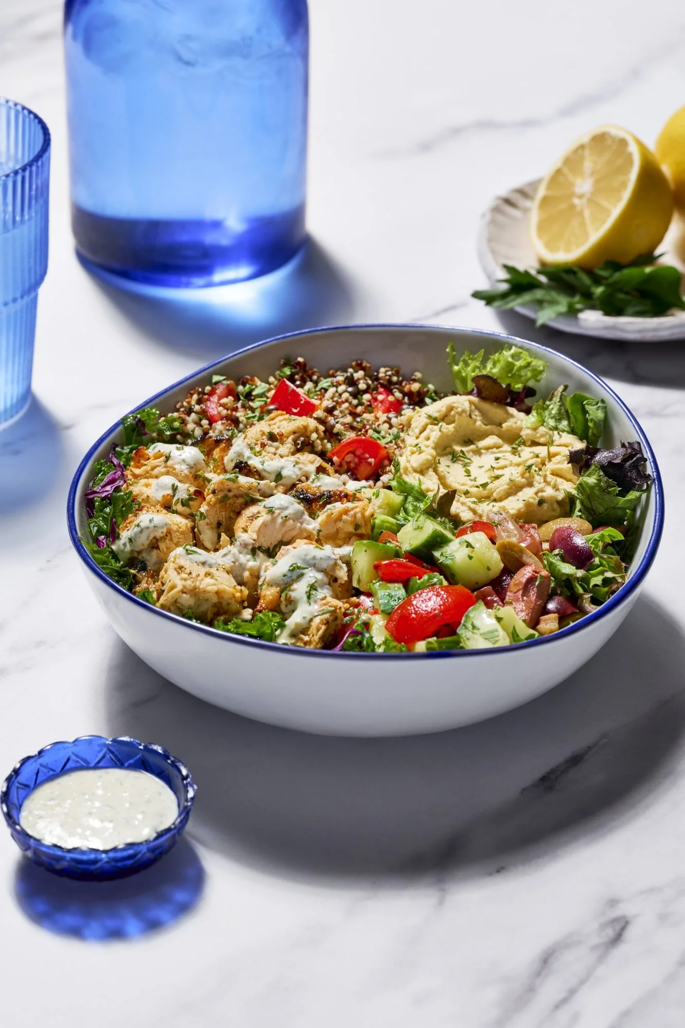 Mediterranean chicken salad bowl with hummus, quinoa, vegetables, herbs, and creamy dressing, served with lemon wedges