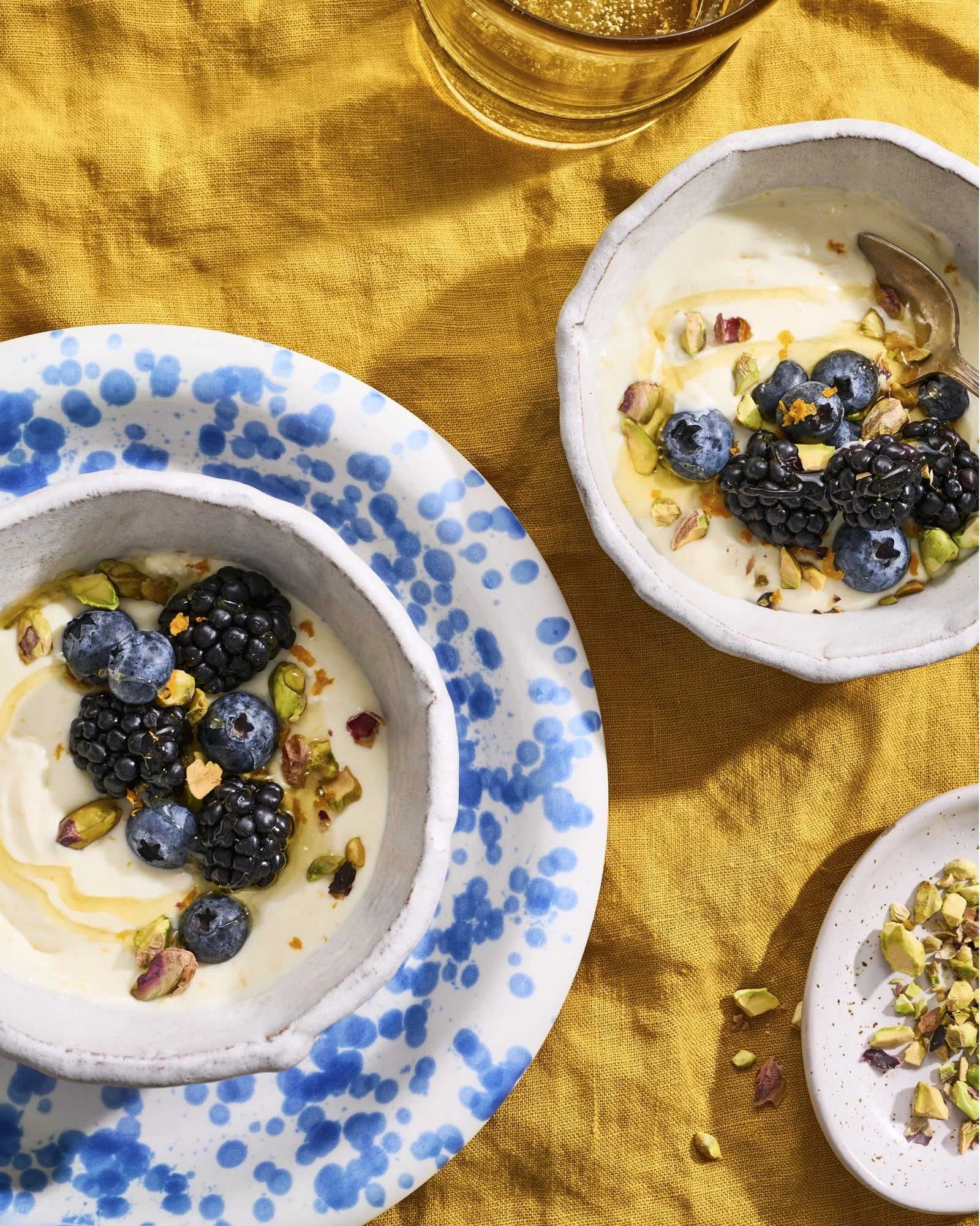Close-up of bowls of creamy yogurt topped with blueberries, blackberries, and chopped pistachios, placed on a yellow tablecloth with scattered pistachio pieces.