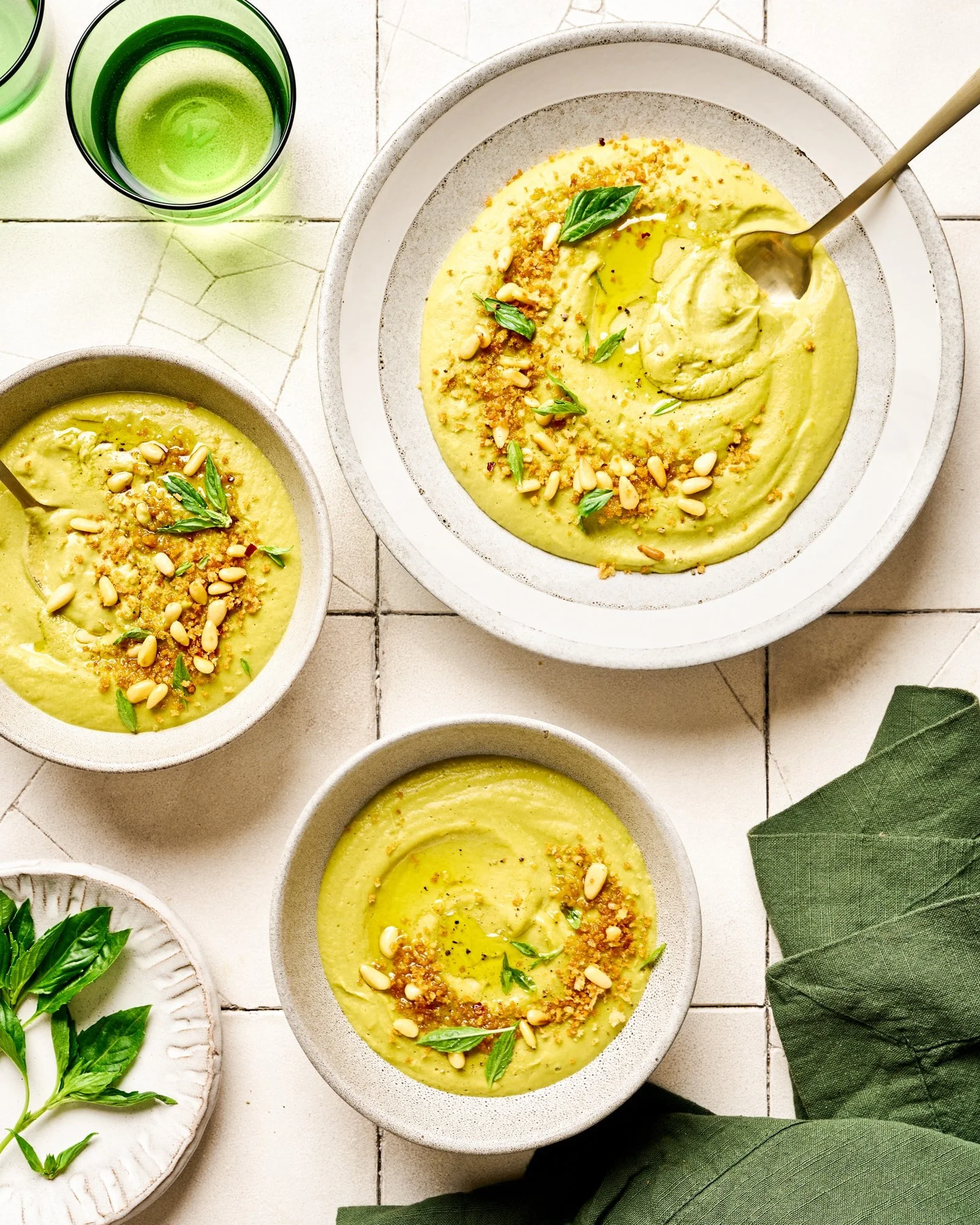 Bowl of creamy green soup garnished with pine nuts, basil leaves, and breadcrumbs on a white plate, with a glass of green beverage on a white tiled table.