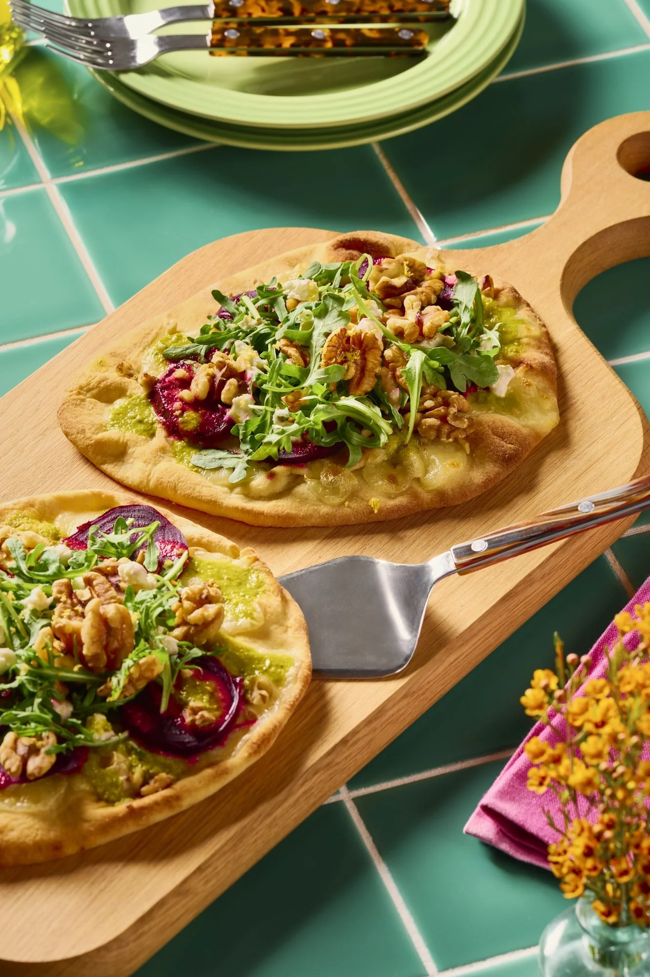 Two flatbreads topped with pesto, roasted beets, arugula, feta cheese, and walnuts on a wooden serving board