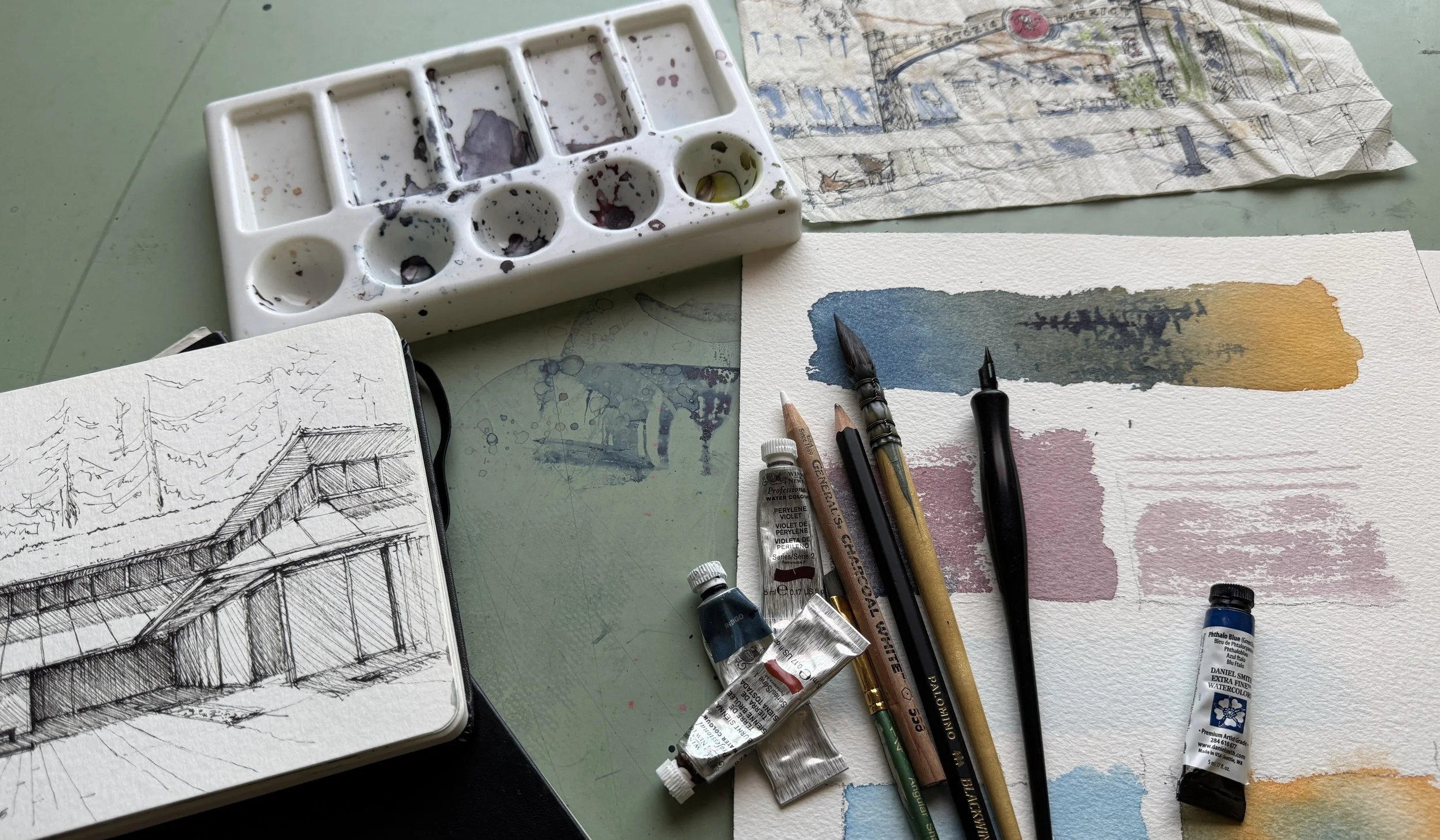 Art supplies including watercolor paints, brushes, ink pens, and a sketchbook displaying a landscape drawing are arranged on a table, with watercolors and tools used for watercolor painting visible.
