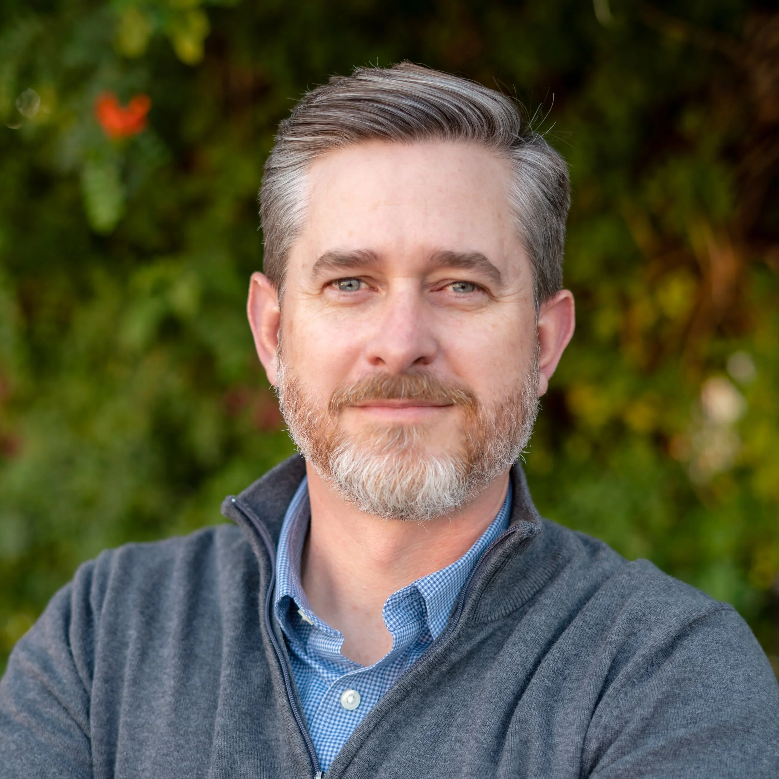 Close-up of a smiling middle-aged man with gray hair, blue eyes, and a beard, wearing a gray zip-up sweater over a blue checkered shirt, outdoors with blurred green foliage background.