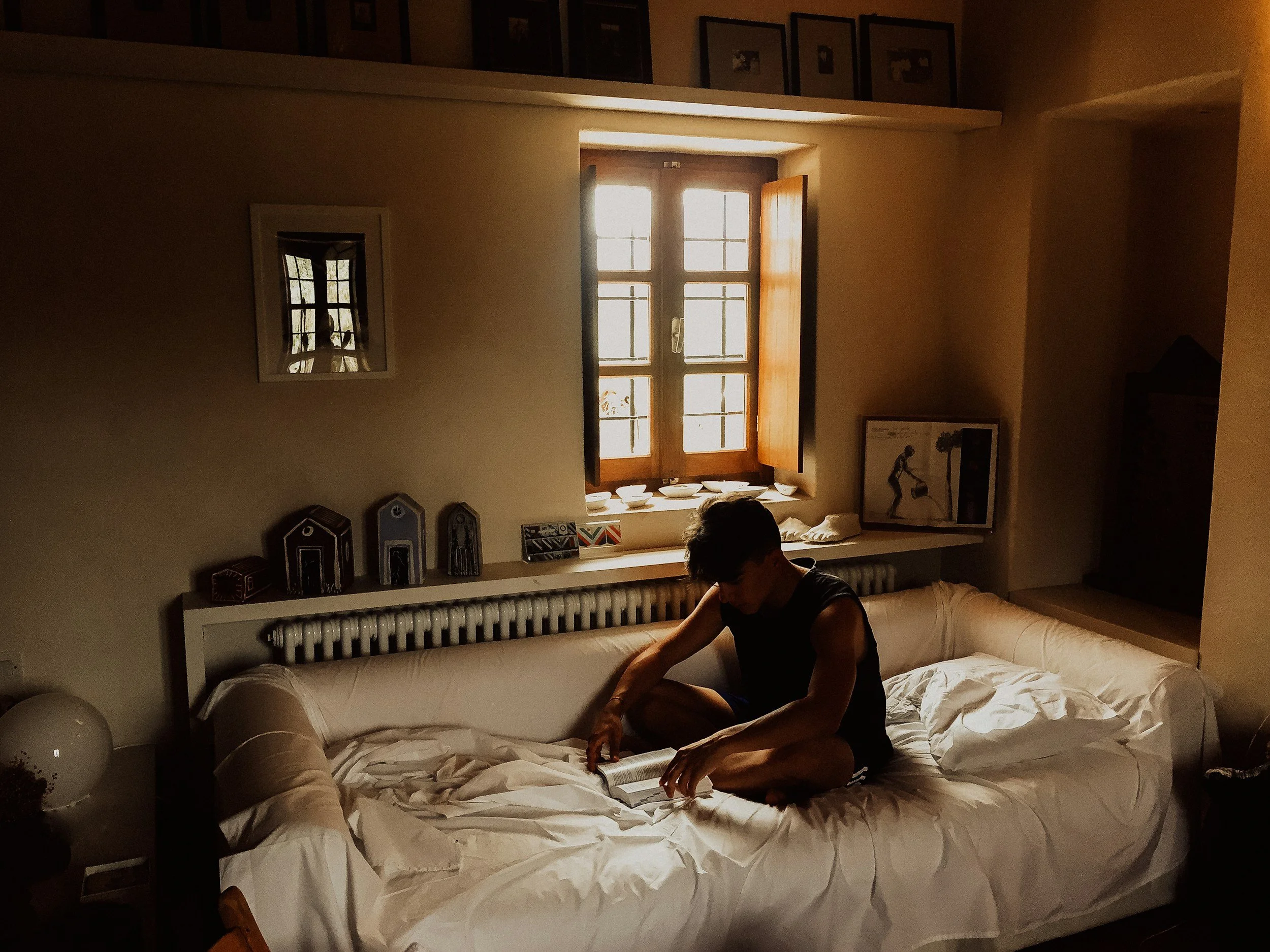 A person sitting on a large bed in a warmly lit room, reading a book with their head bowed down. The room has a window with wooden frames and open shutters, decorative framed pictures on the walls, and various objects on the windowsill and shelves.