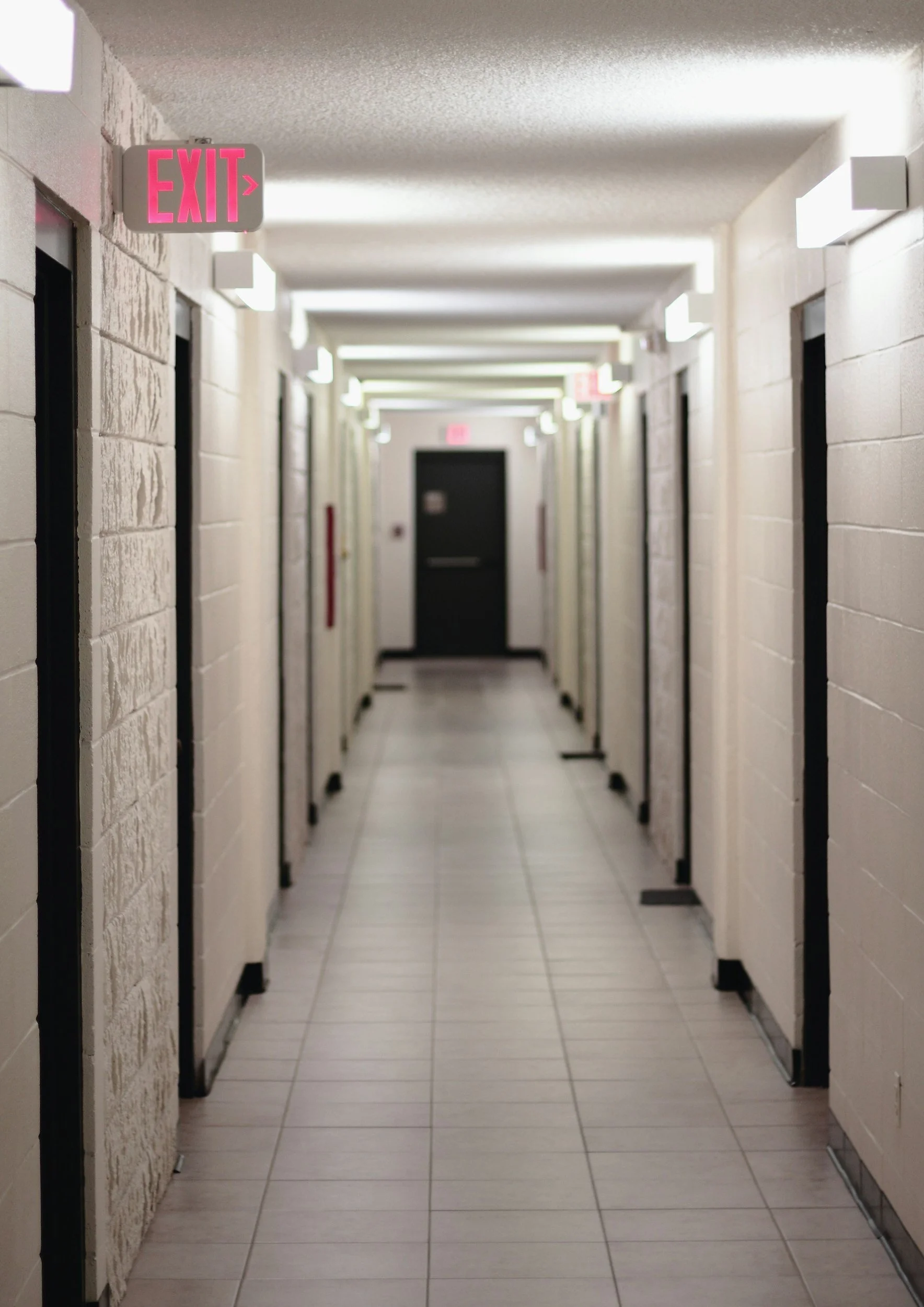 A hallway with multiple elevator doors on both sides, white brick walls, and tiled floor. There is an illuminated pink 'EXIT' sign with an arrow pointing to the right at the beginning of the corridor.