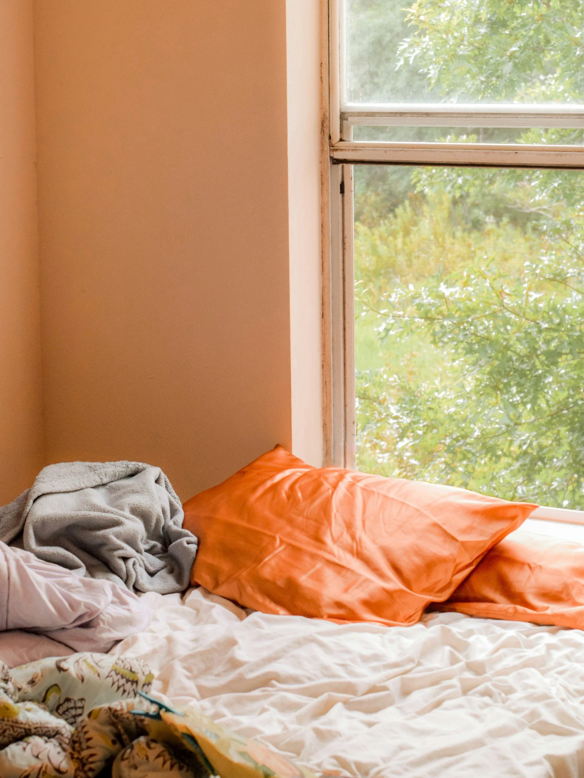 A bed with orange pillows and white sheets next to a window with green trees outside, sunlight coming through.