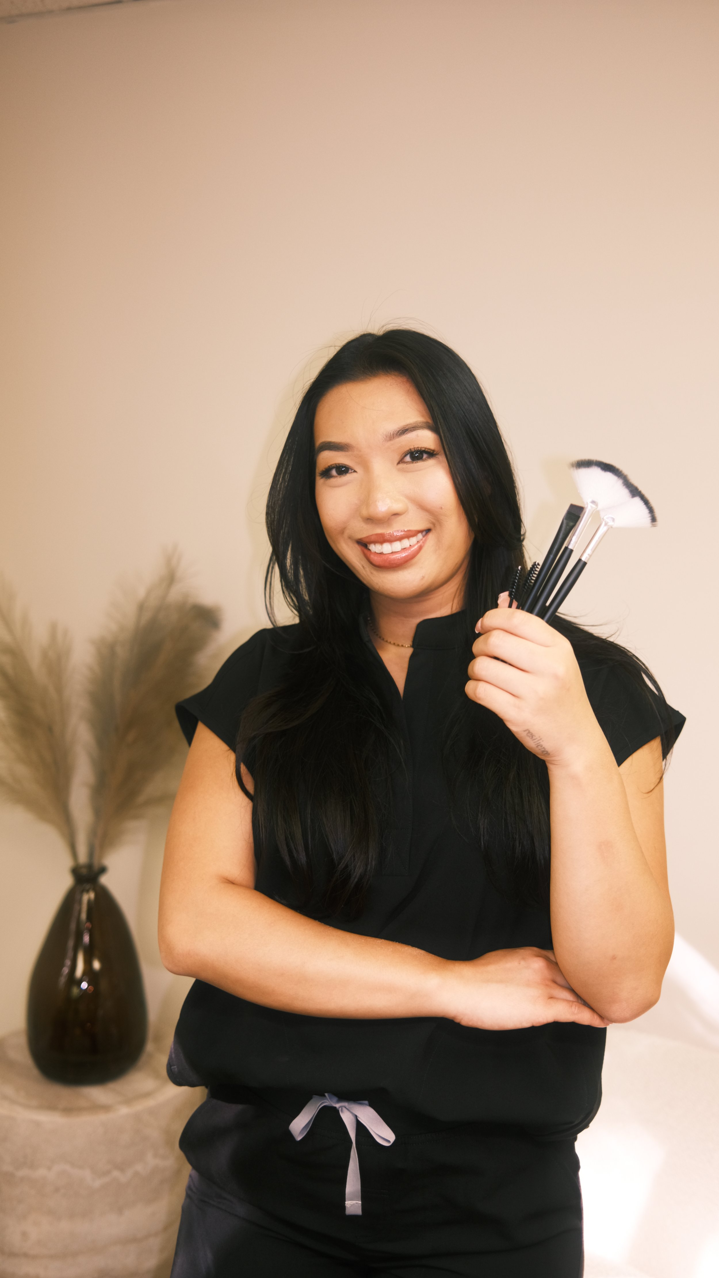 A woman smiling and holding makeup brushes, standing in a room with a beige wall and a brown vase with decorative dried plants in the background.