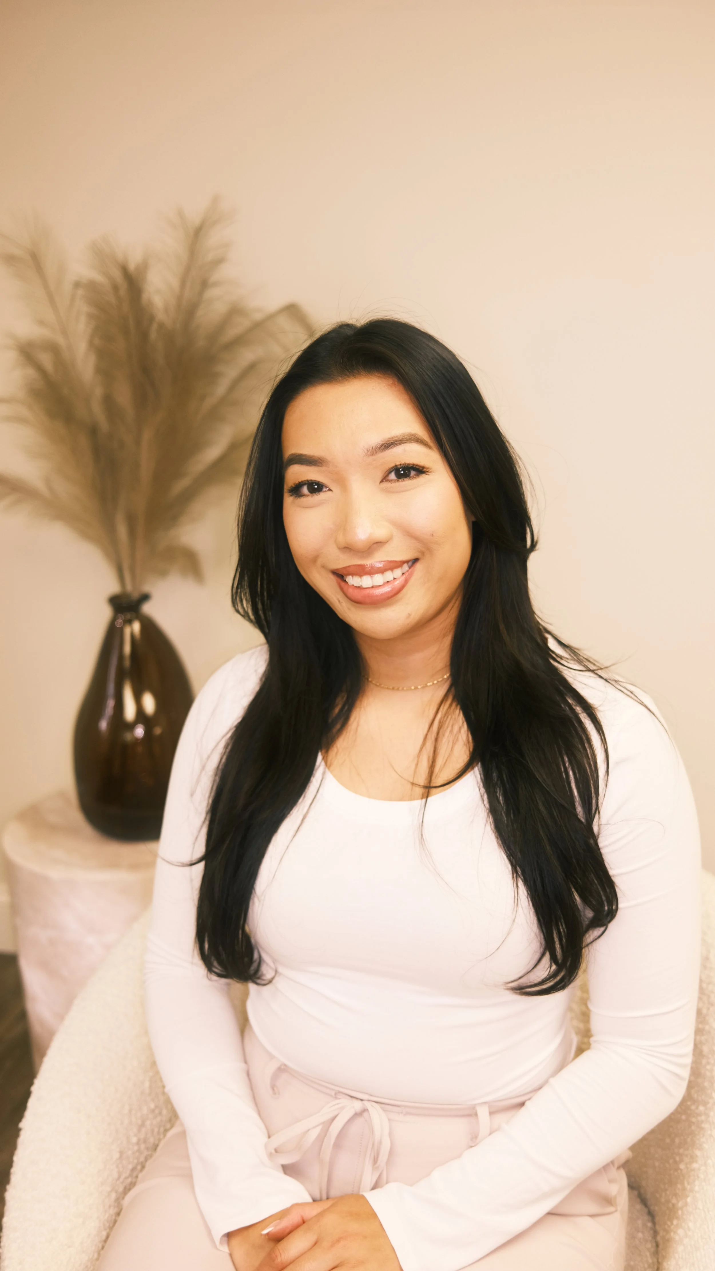 A woman with long black hair and a white top sitting on a cream-colored chair next to a side table with a brown vase containing pampas grass, smiling at the camera.