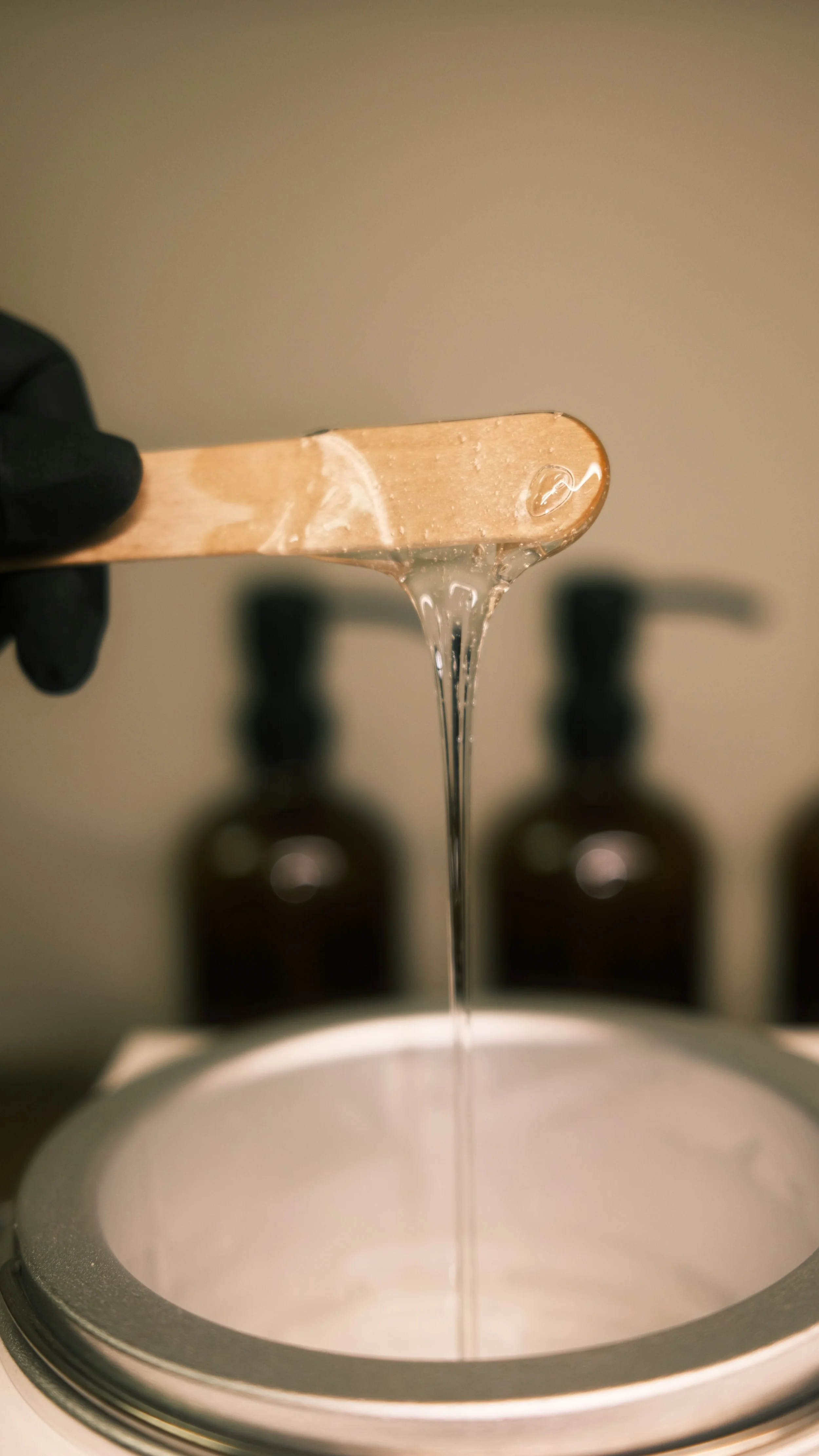 A close-up of a wooden spoon with clear viscous liquid dripping into a container. Two dark bottles blurred in the background.
