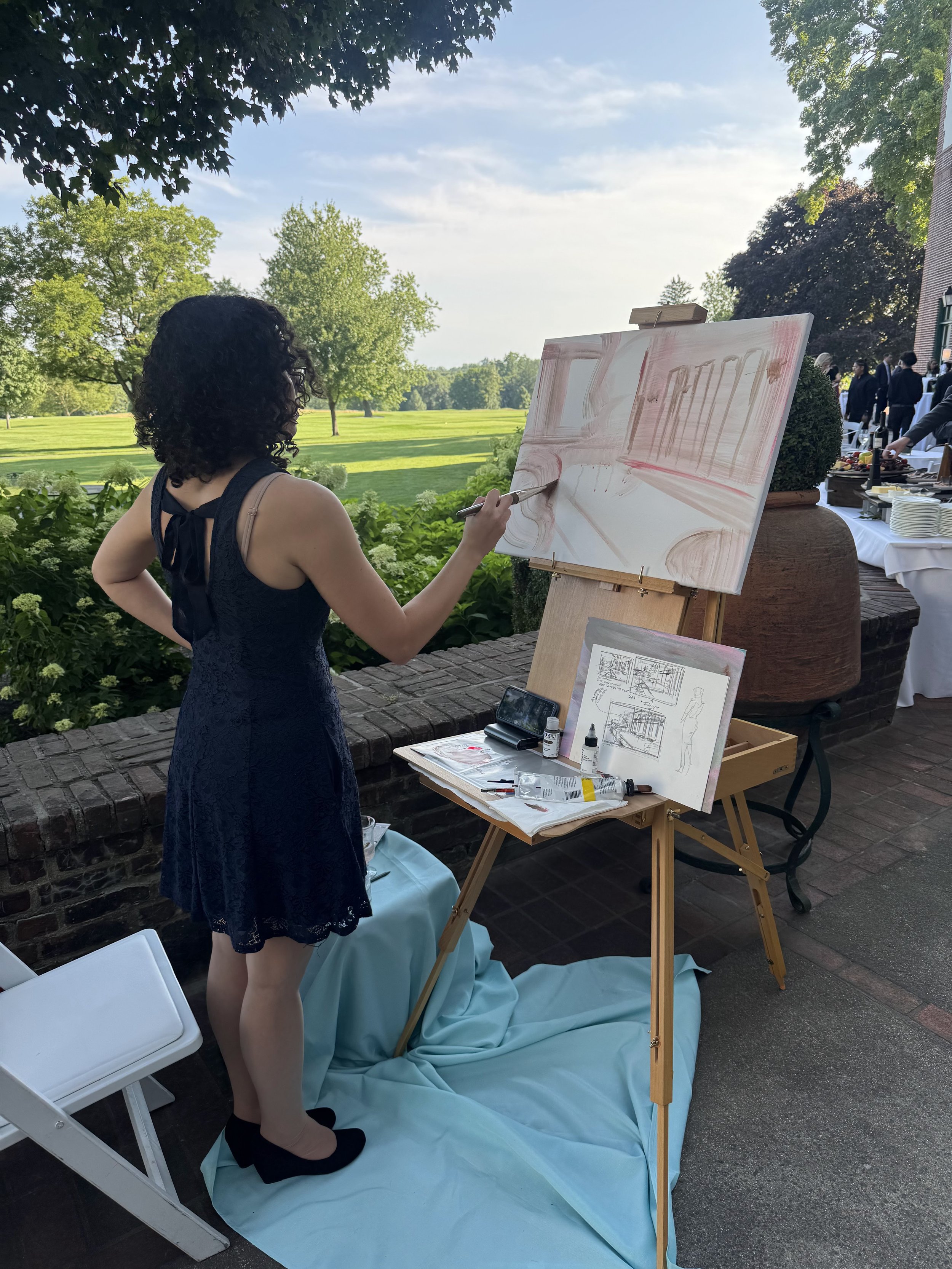 A woman in a navy blue dress is painting on a large canvas set on an easel outdoors under trees on a bright, sunny day. There is a table with art supplies and sketches nearby, and a gathered group of people in the background.