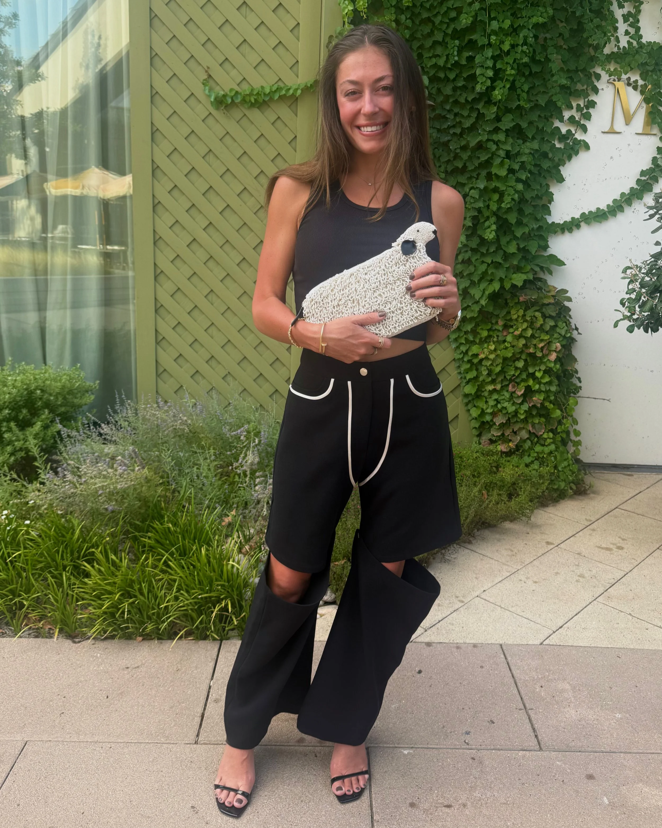 A smiling woman in a black sleeveless top and black wide-leg pants holding a decorative white object, standing on a sidewalk in front of greenery and a building with a green lattice and white wall with green ivy.