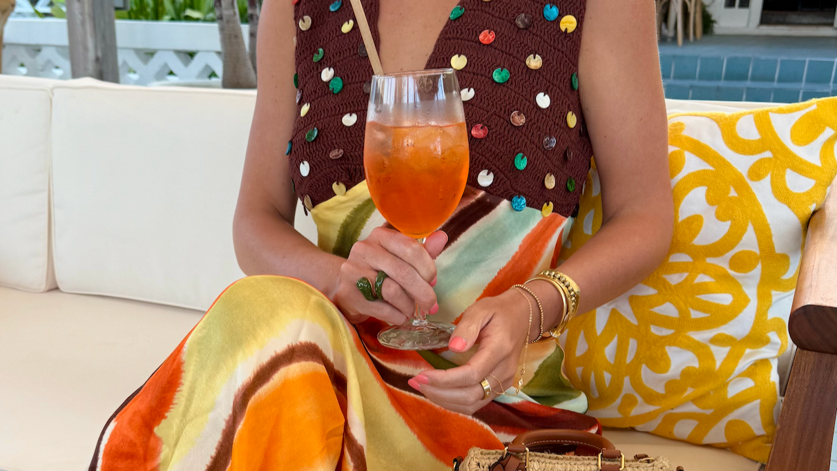 A woman in colorful clothing sitting on a white outdoor couch, holding an orange-colored drink in a wine glass. She is wearing jewelry and sitting next to a yellow patterned pillow.