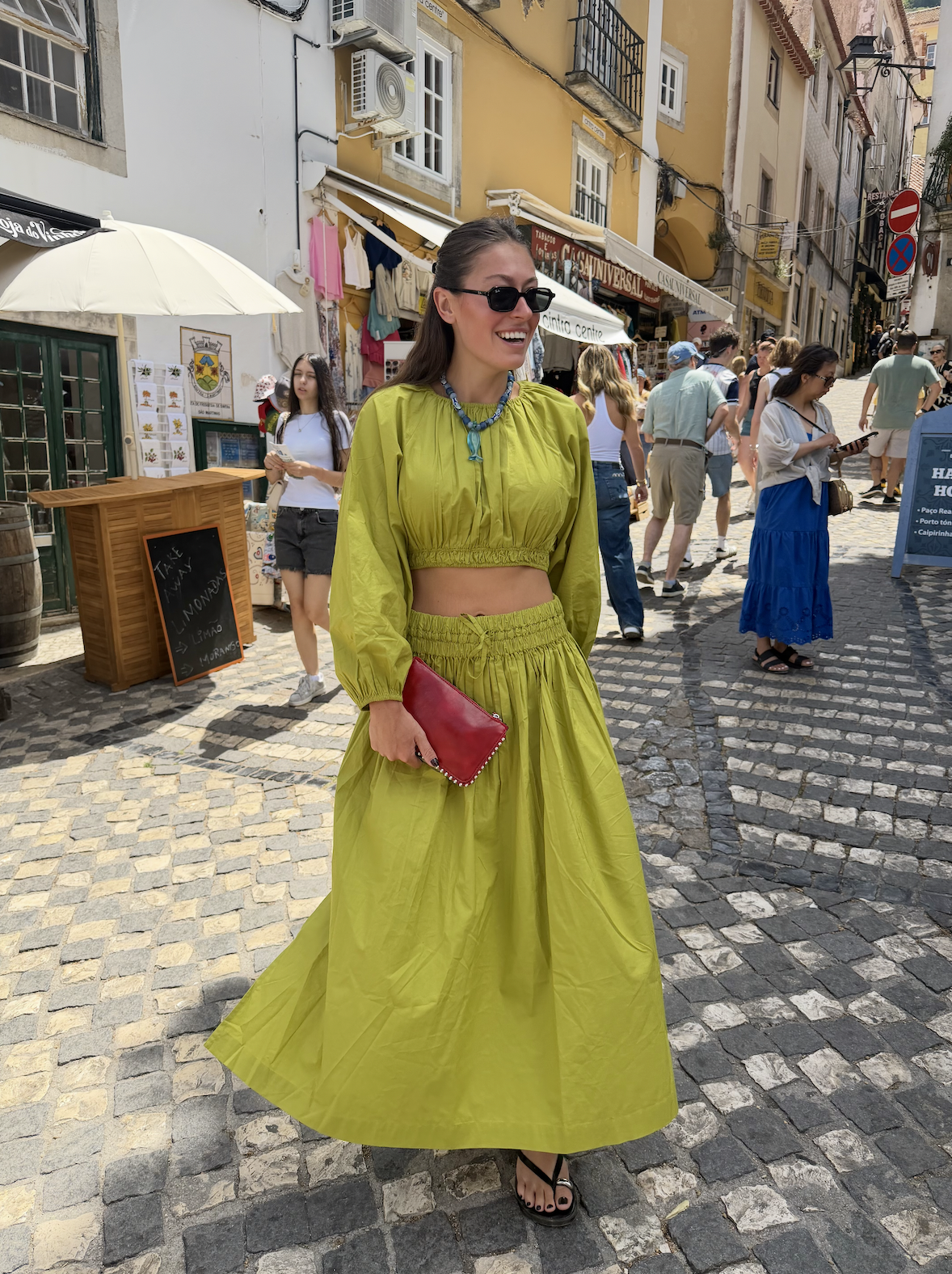 A smiling woman dressed in a bright yellow crop top and matching long skirt holding a red clutch purse walking on a cobblestone street in a busy outdoor market area with colorful buildings and shops.