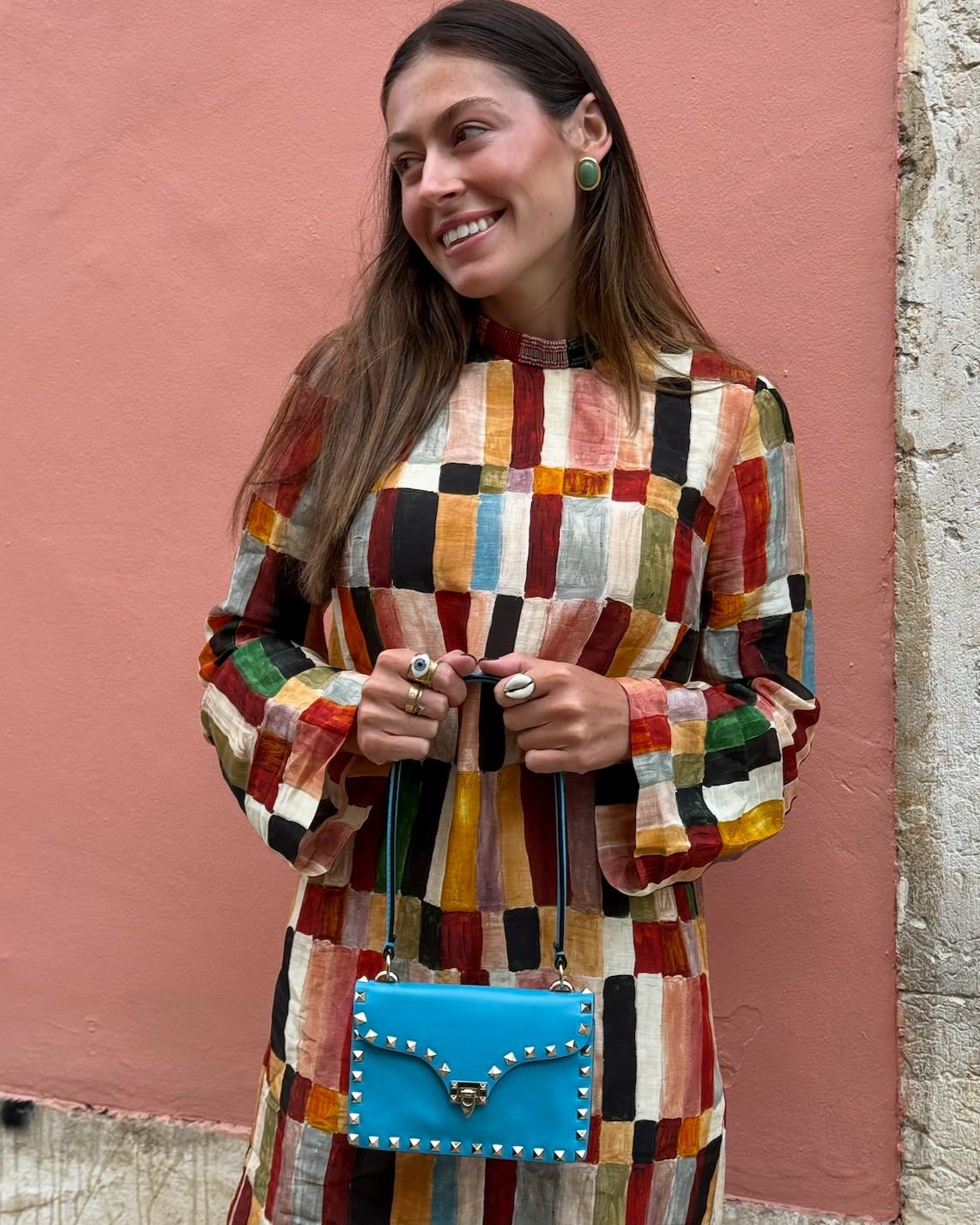 A woman wearing a colorful patchwork dress holding a small blue purse with silver studs, standing in front of a pink wall with a textured section on the right.