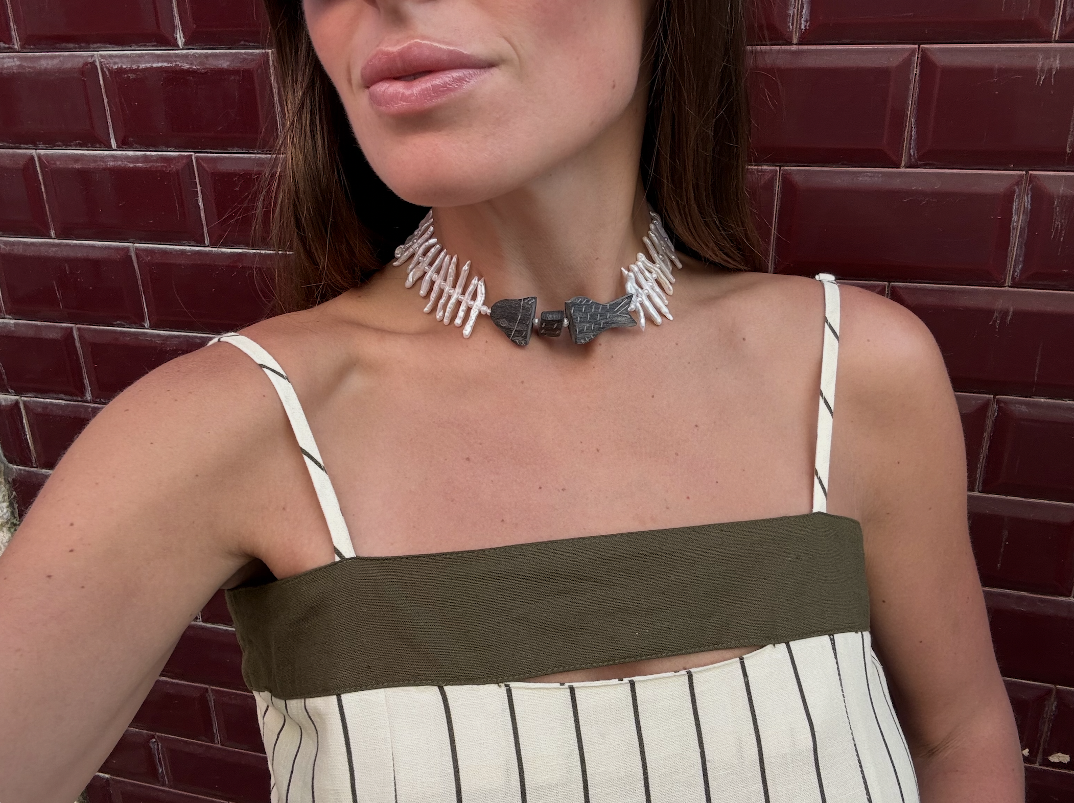 Close-up of a woman wearing a necklace made of white shell-like beads and a large black and gray bow-shaped pendant. She is wearing a top with white and black striped straps, standing in front of a red brick wall.