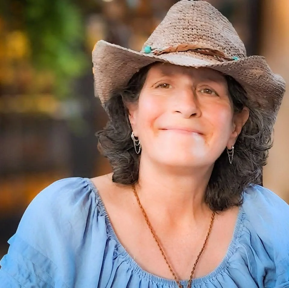 A woman outdoors wearing a cowboy hat and a blue blouse, smiling.
