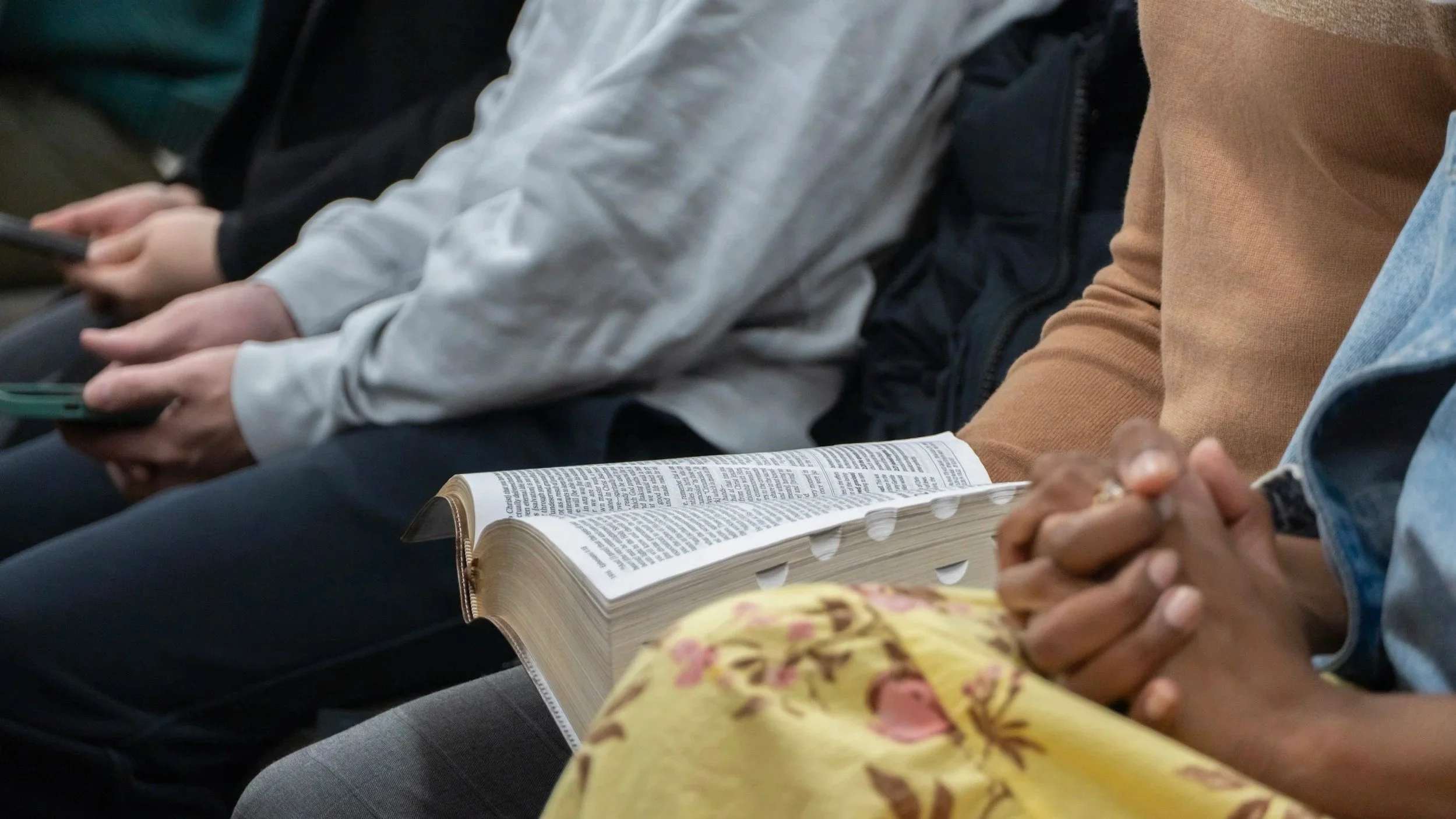 People seated in a row, holding their hands together, with one person reading a large Bible, another person using a mobile phone, and others sitting quietly.