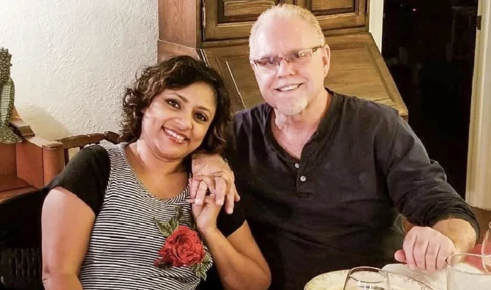 A couple smiling and holding hands at a dining table, with a woman of South Asian descent and a man of Caucasian descent, in a cozy indoor setting.