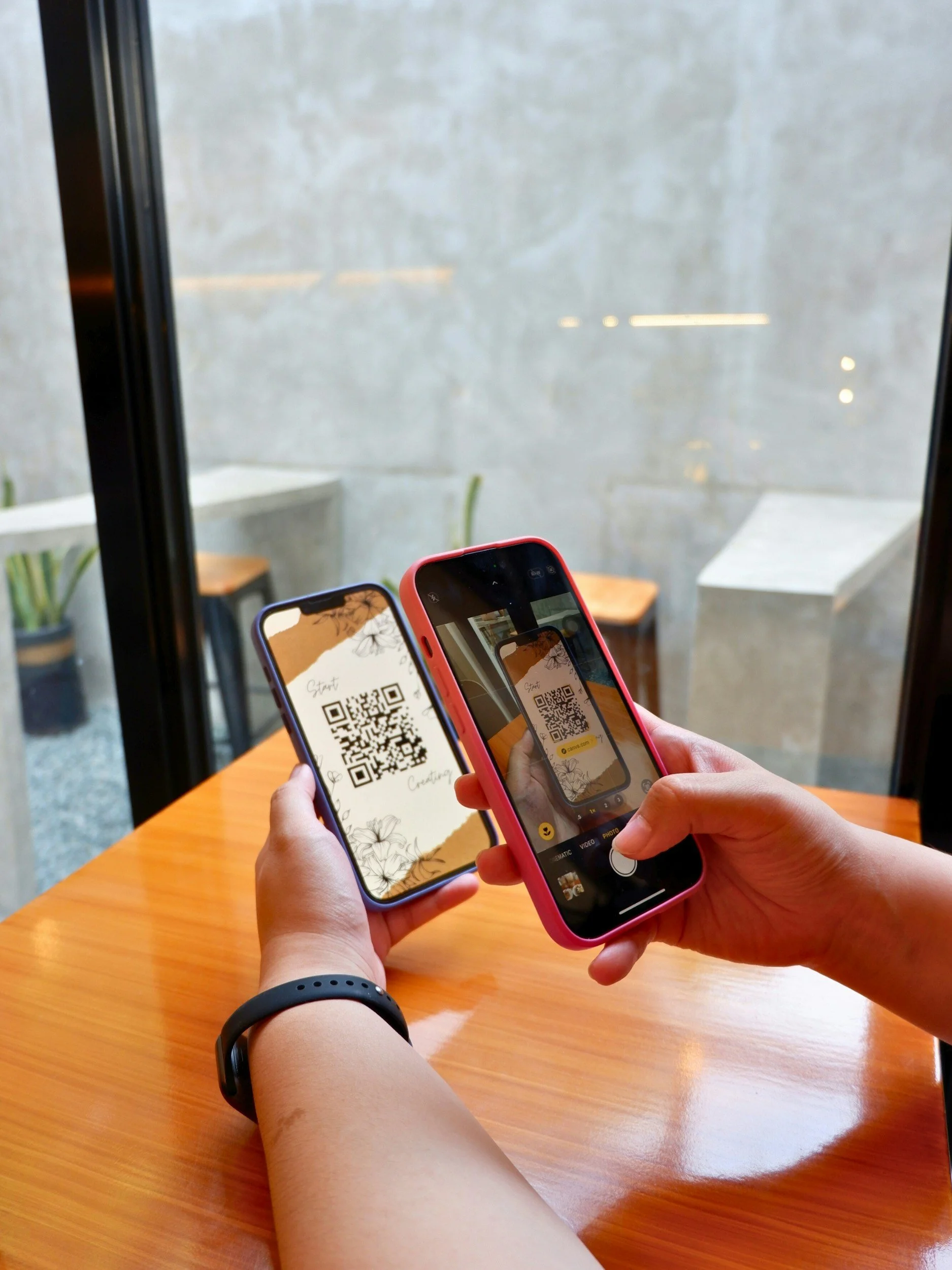 Person holding two smartphones with QR codes on the screens, sitting at a wooden table in a cafe, with a view of two chairs and a concrete wall outside the window.