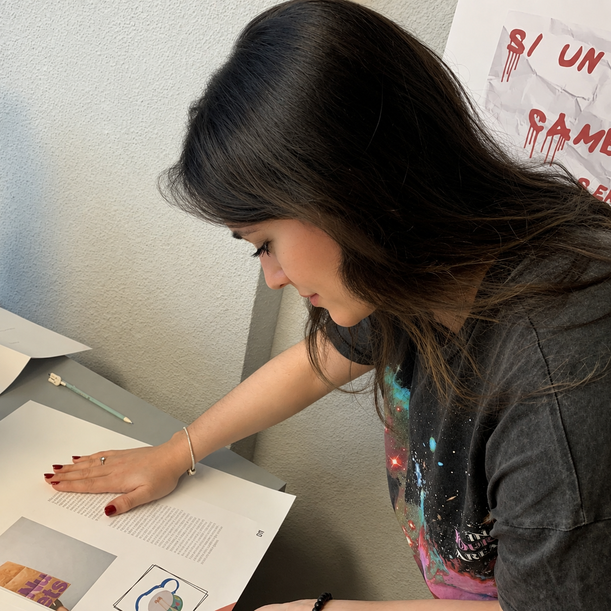 A young woman with long, dark brown hair is reading a printed page on a table. She has a silver bracelet on her left wrist, red nail polish, and appears focused on the material. There is a pencil and some paper nearby, and a poster with red text on a wrinkled white background is partially visible on the wall.