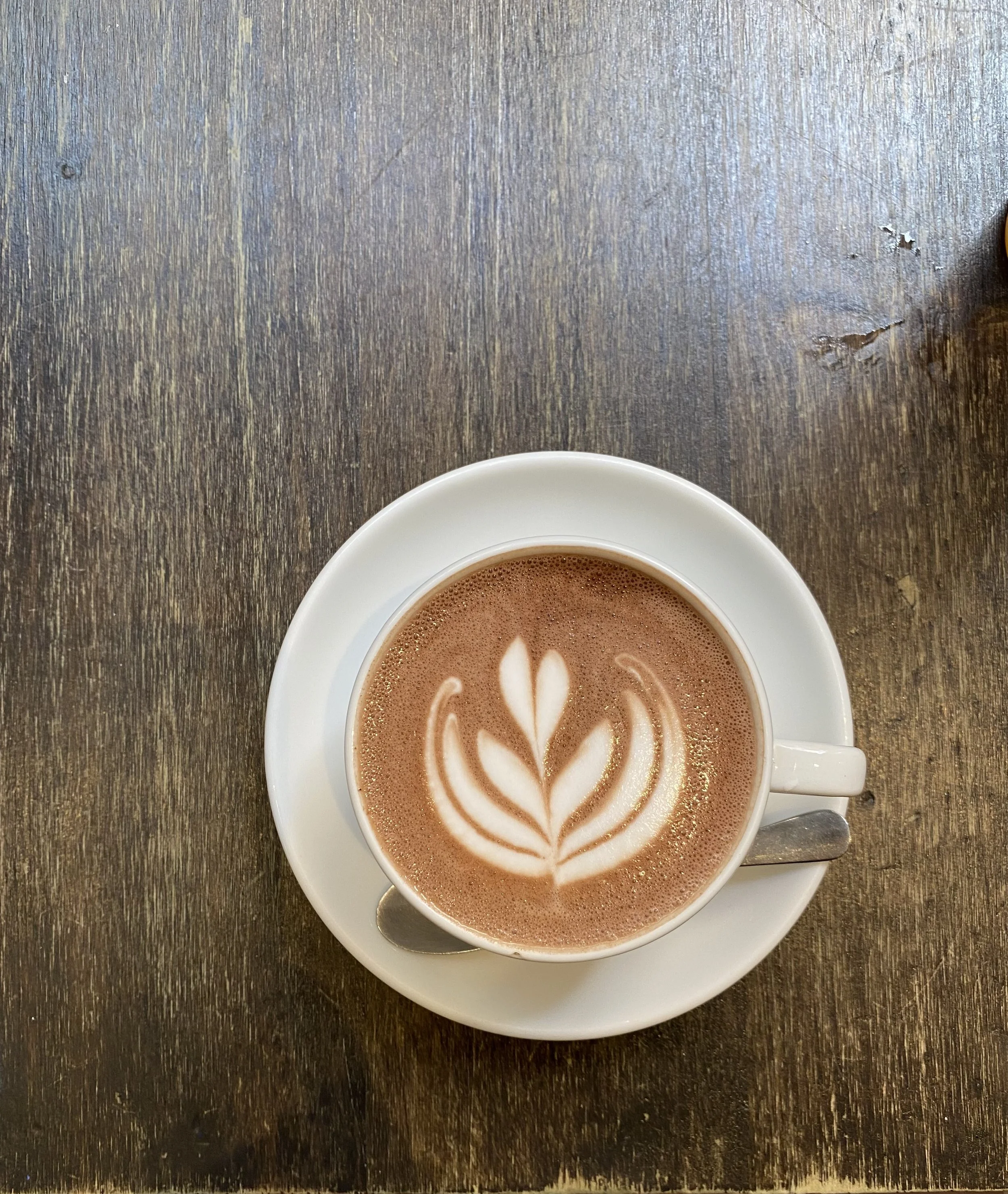 A white cup of hot chocolate with latte art, sitting on a white saucer with a spoon on a wooden table.