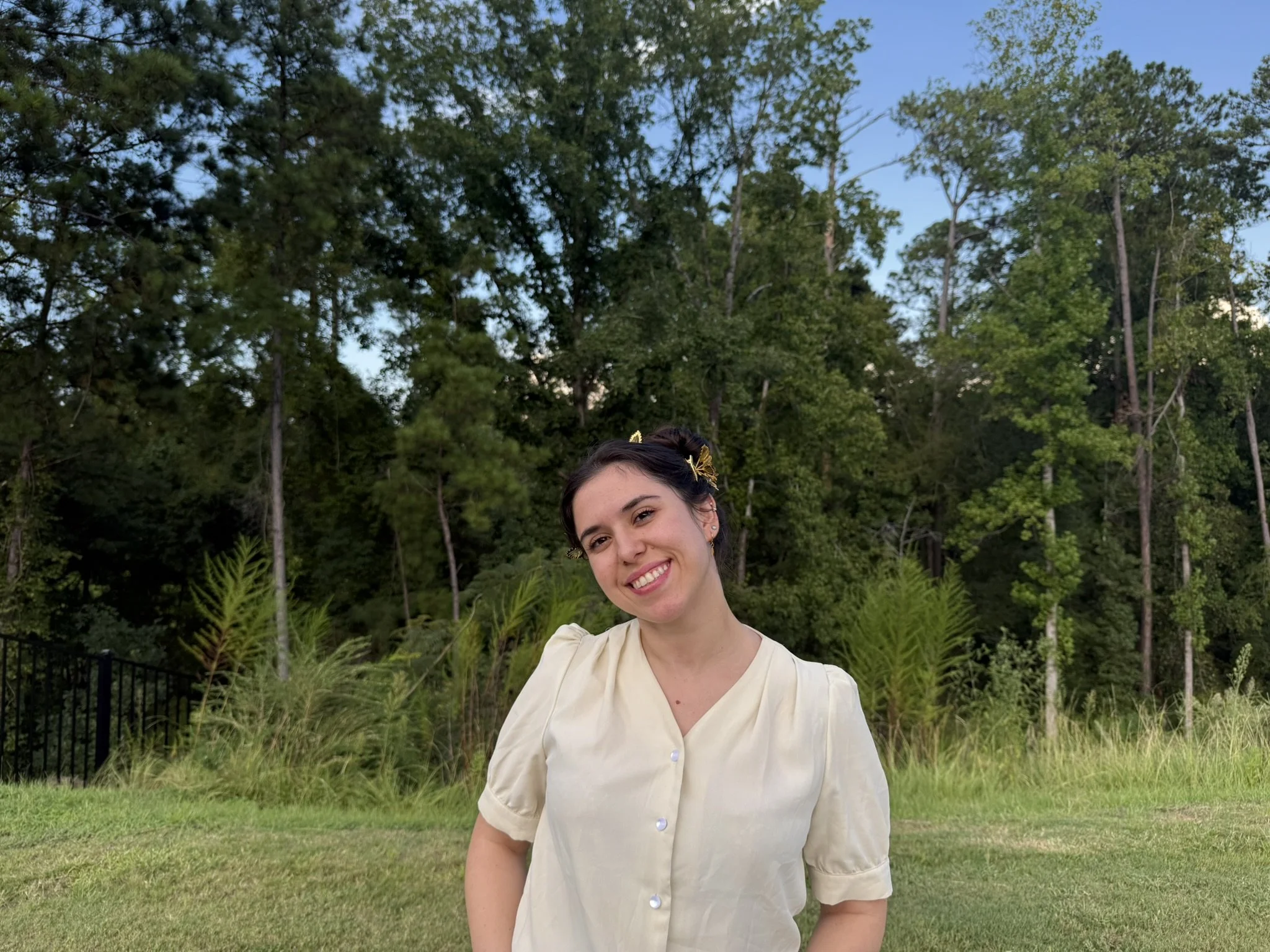 A woman with dark hair, wearing a light-colored overall dress, smiling and standing outdoors in front of tall trees and bushes.