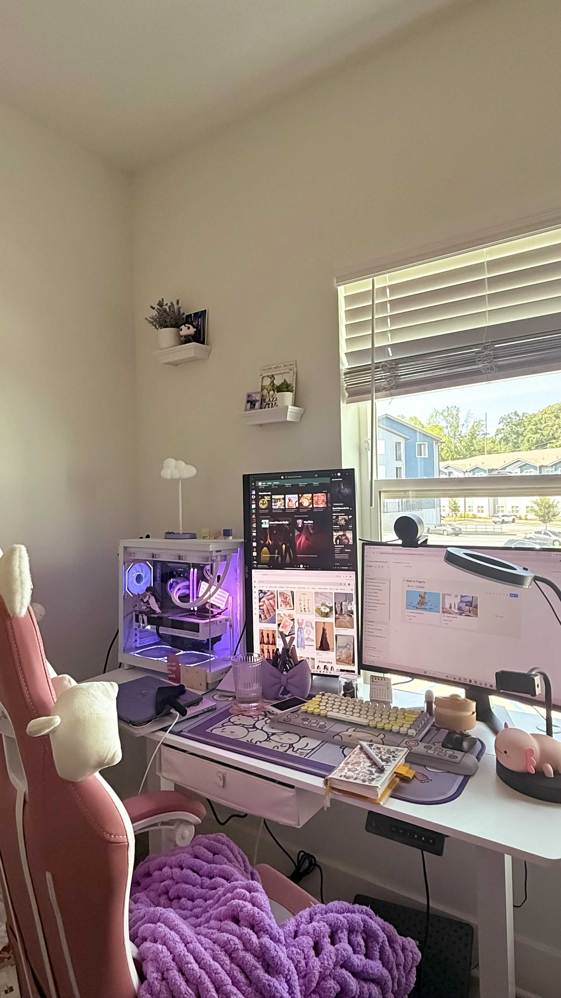 A workspace with a desk, pink chair, plush blanket, multiple monitors, a white desk lamp, and a computer case with purple lighting near a window with blinds, and two shelves with plants and decorations on the wall.