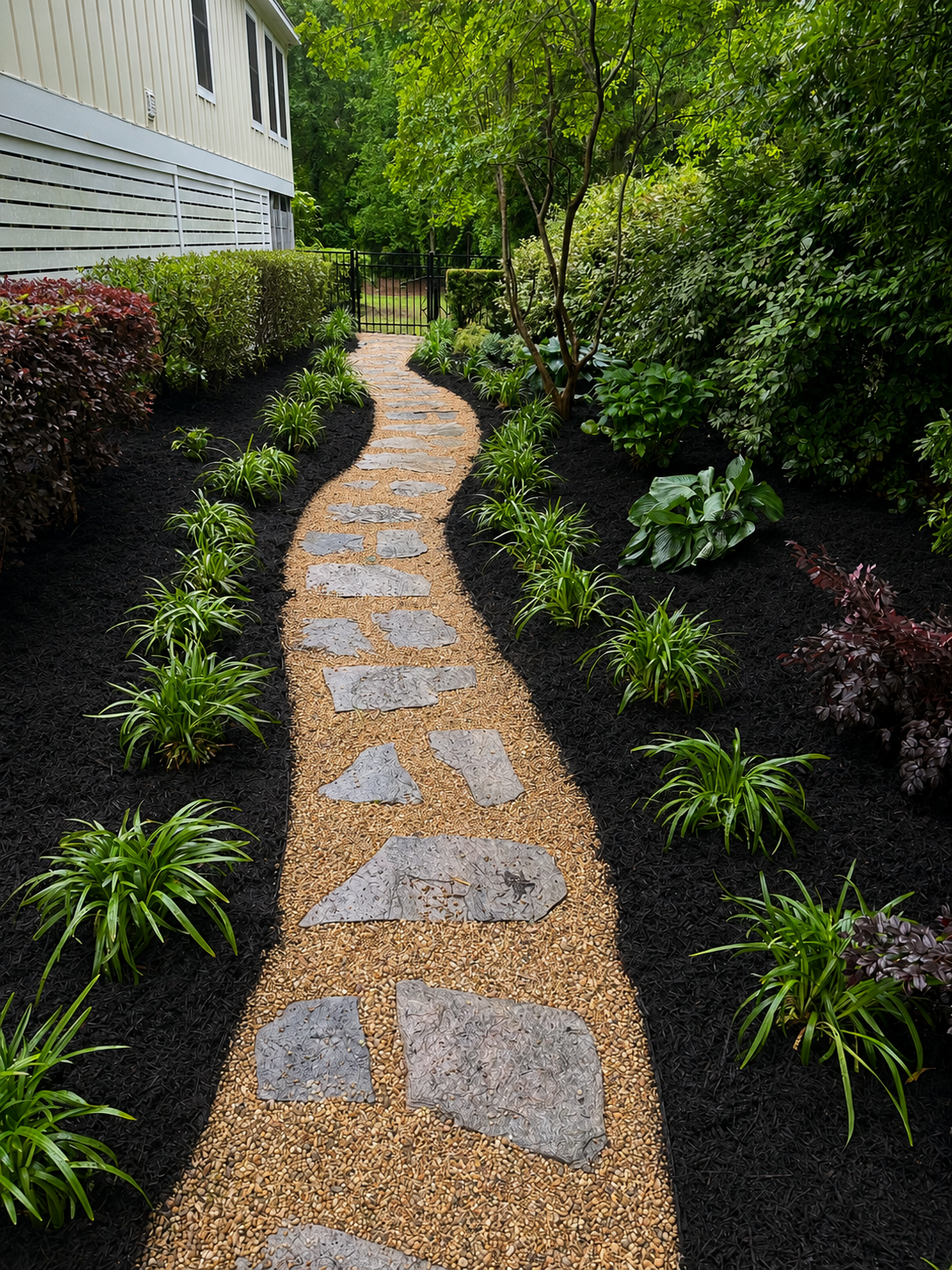A narrow garden pathway with stone steps and gravel, bordered by green plants and shrubs, leading to a black gate with trees in the background.
