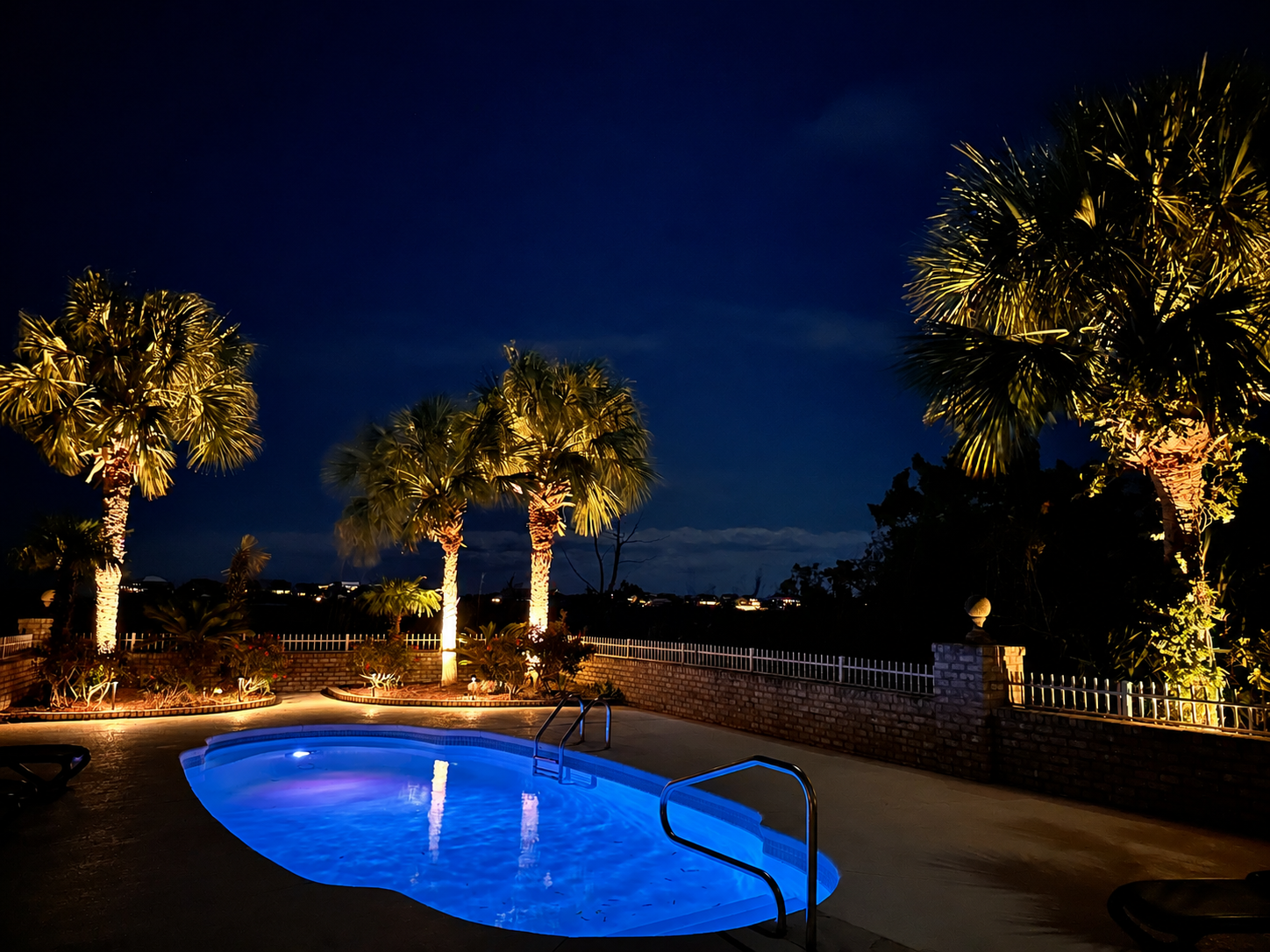 Night view of a lit outdoor swimming pool with illuminated palm trees and a brick wall with a metal fence in the background.