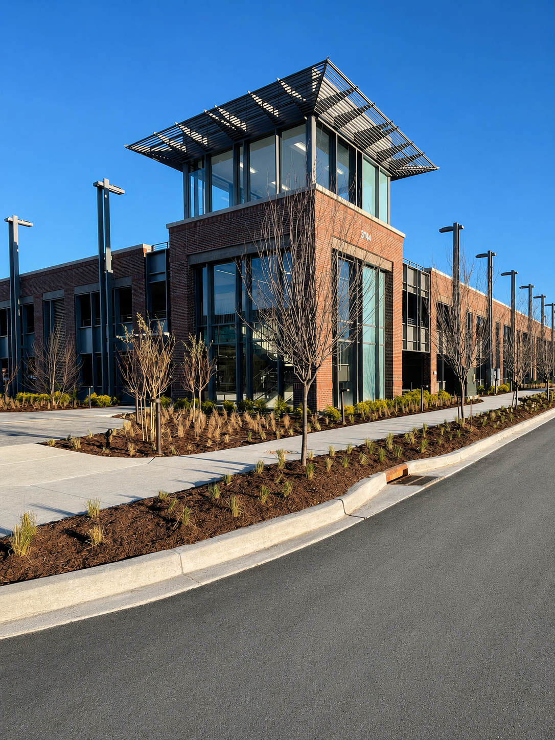 Modern commercial building with brick and glass facade, surrounded by landscaped area with small trees and shrubs, clear blue sky.