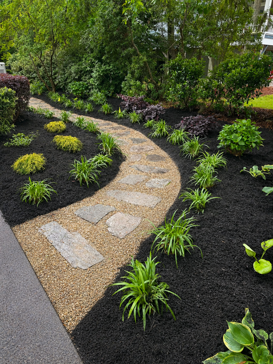 A garden with a winding stone pathway surrounded by lush green plants and bushes, with dark mulch covering the ground.