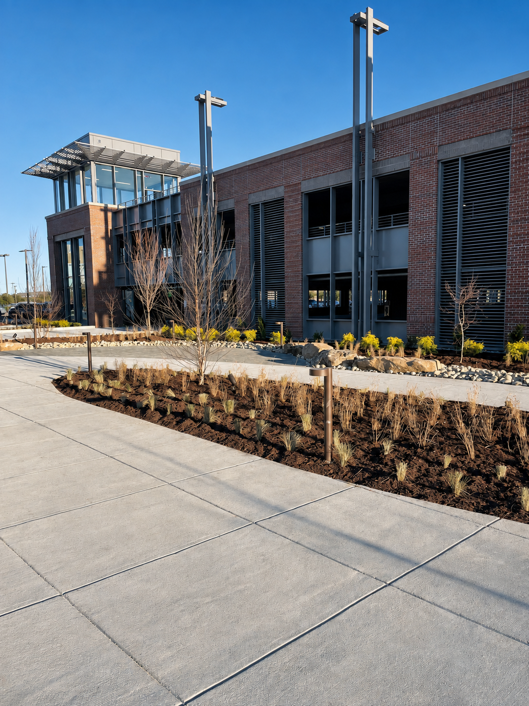 Modern building with brick and metal exterior, landscaped sidewalk with small trees and plants, under a clear blue sky.