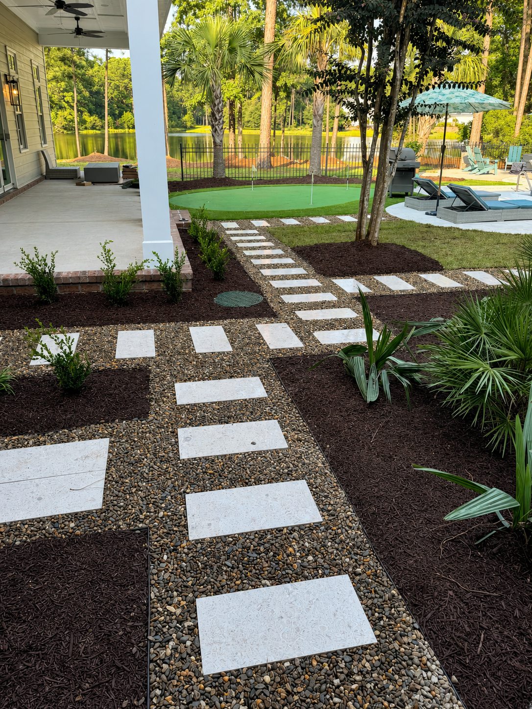 A backyard patio area with a white wooden porch on the left, a stone pathway with white stepping stones and gravel, landscaped plants, and a play putting green near a lake surrounded by trees in the background. There are lounge chairs, an umbrella, a