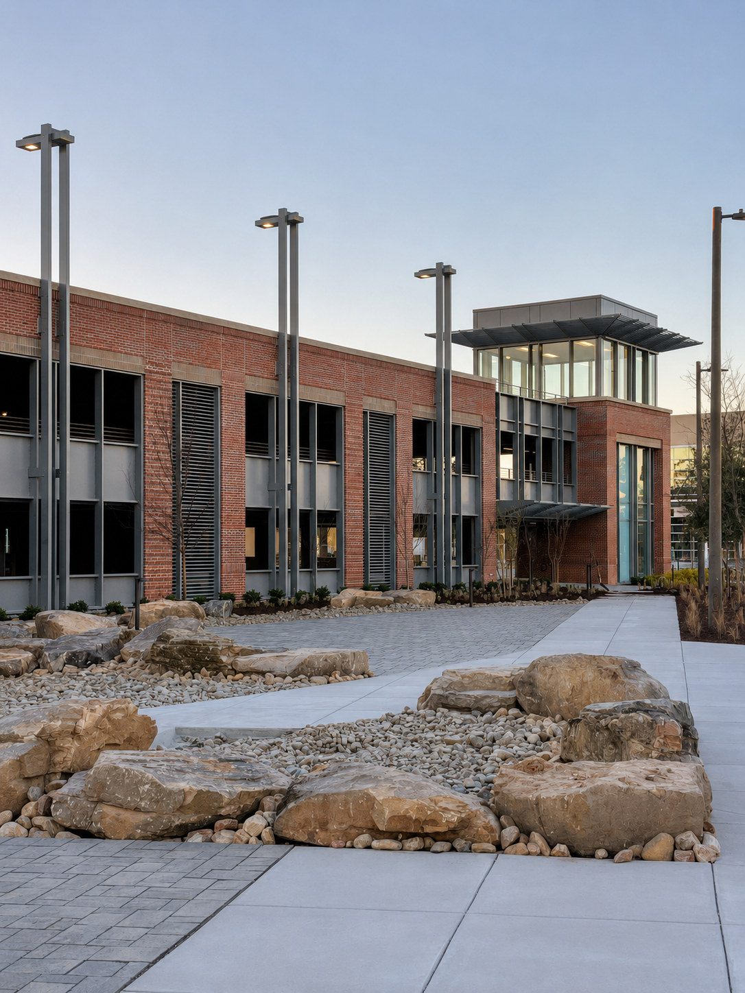 Modern building with red brick and glass windows, surrounded by landscaped rocks and concrete walkways, with tall street lamps