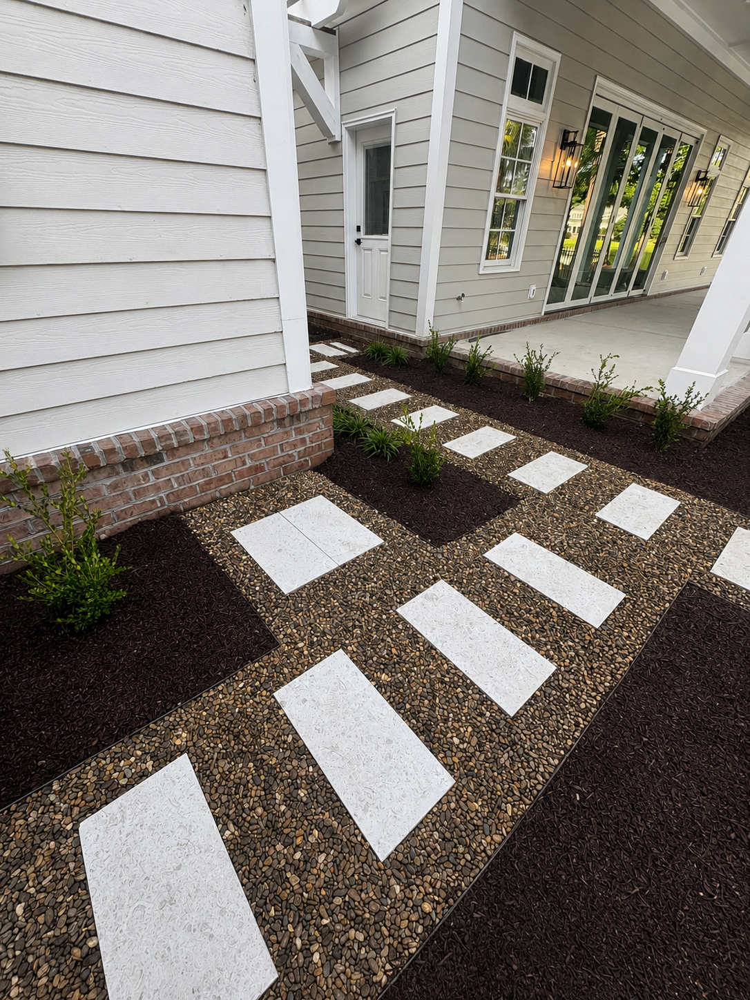 Exterior view of a house with a pathway made of large white tiles set in a pebble surface, bordered by dark mulch and small green plants, next to a patio with a sliding glass door and windows, and wall-mounted outdoor lighting.
