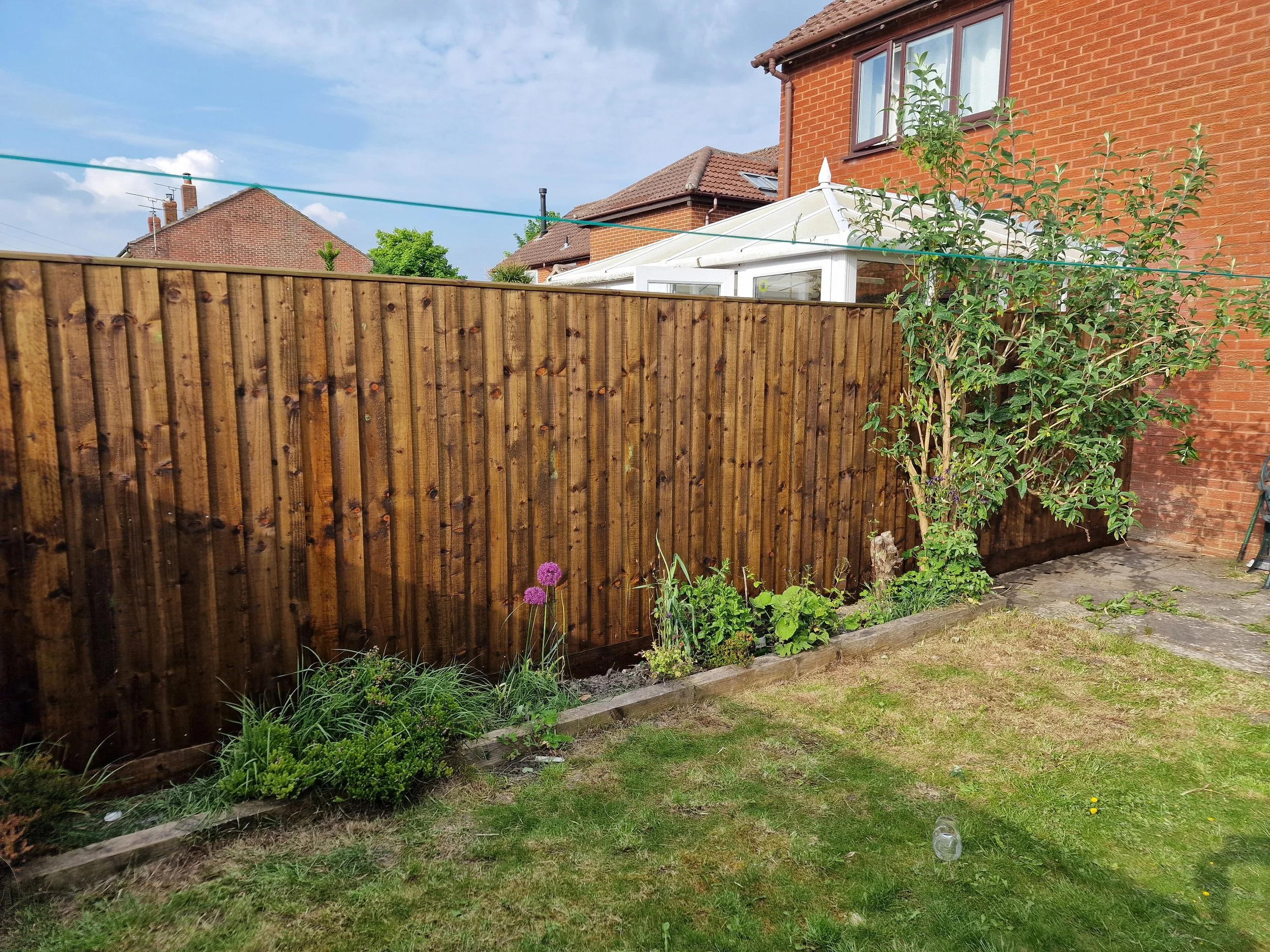A backyard with a wooden fence, some flowers and green grass, and a brick house with a conservatory extension. A clothesline runs across the sky which is partly cloudy.