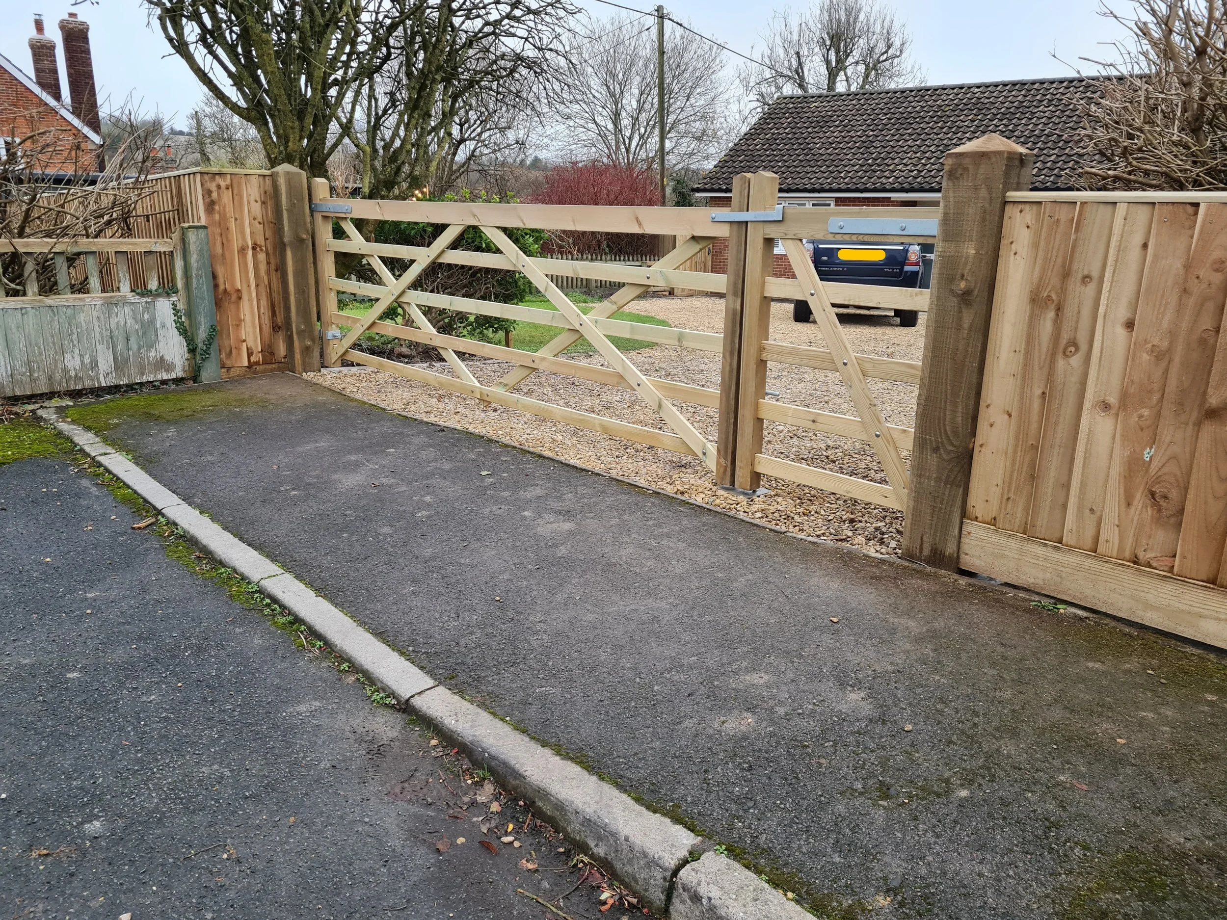 A newly constructed wooden gate and fence in a residential driveway, with a small patch of moss on the edge of the driveway, and a black car parked behind the gate. There are leafless trees and neighboring houses in the background.