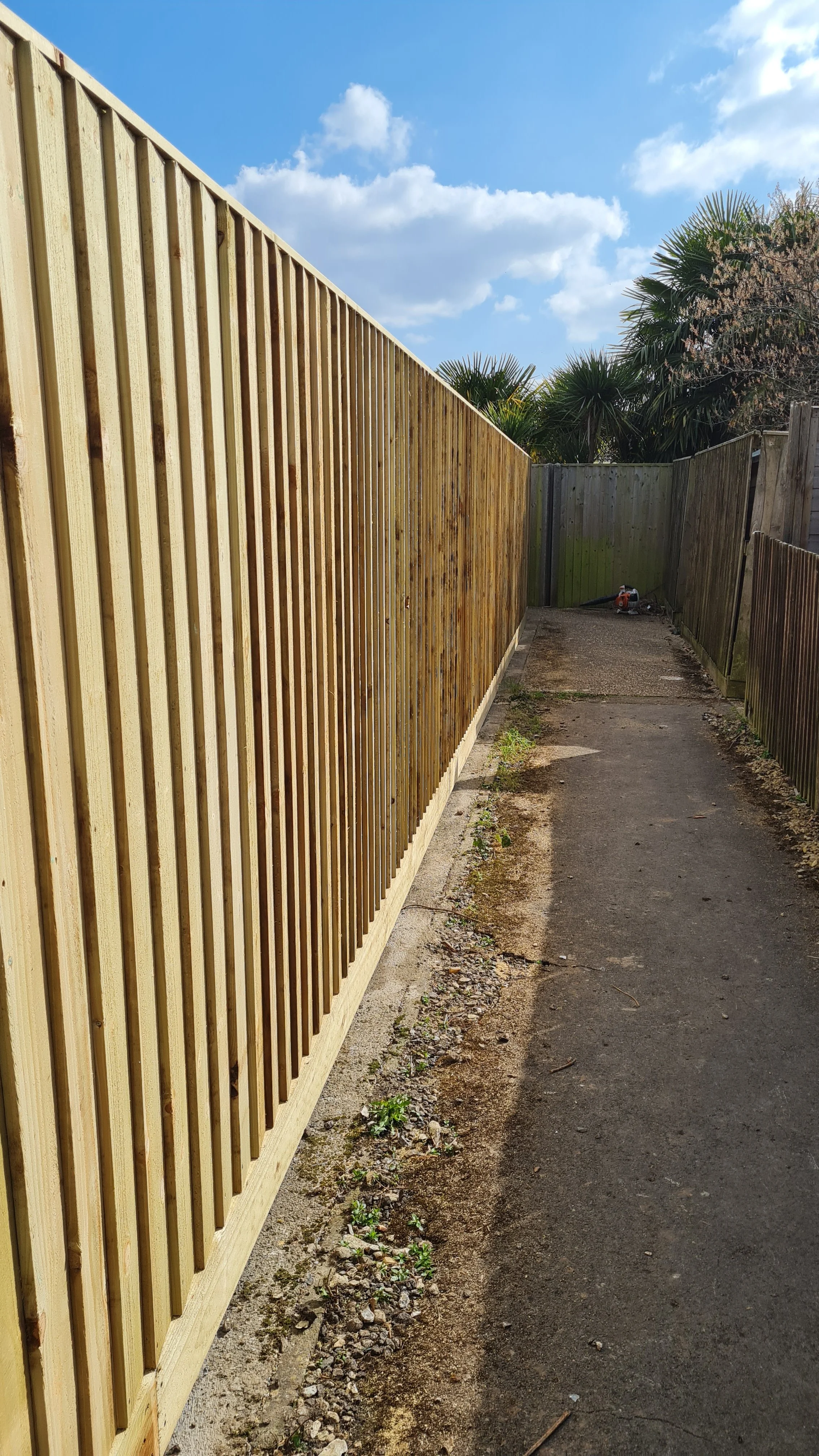 A footpath with wooden fences on either side, a chainsaw near the back on the right, and trees and a partly cloudy sky in the background.