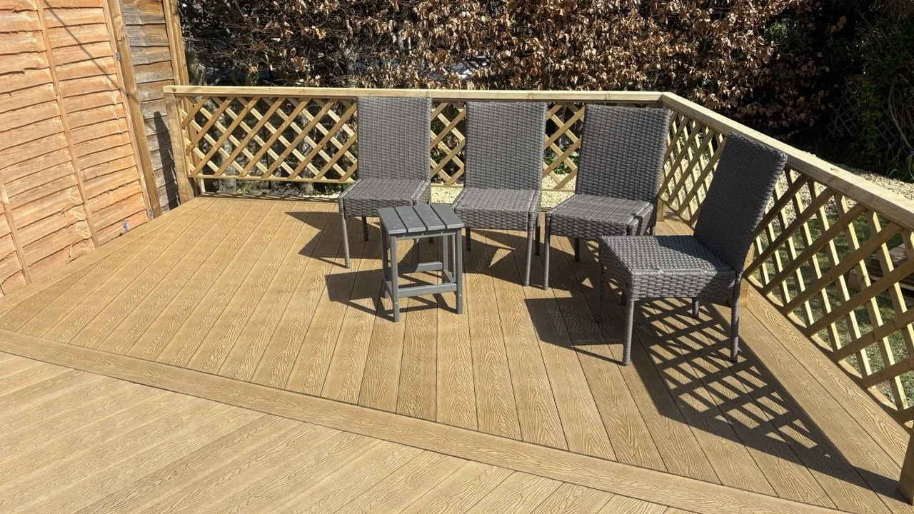 An outdoor wooden deck with four rattan chairs and a small matching table, surrounded by wooden fence and greenery.