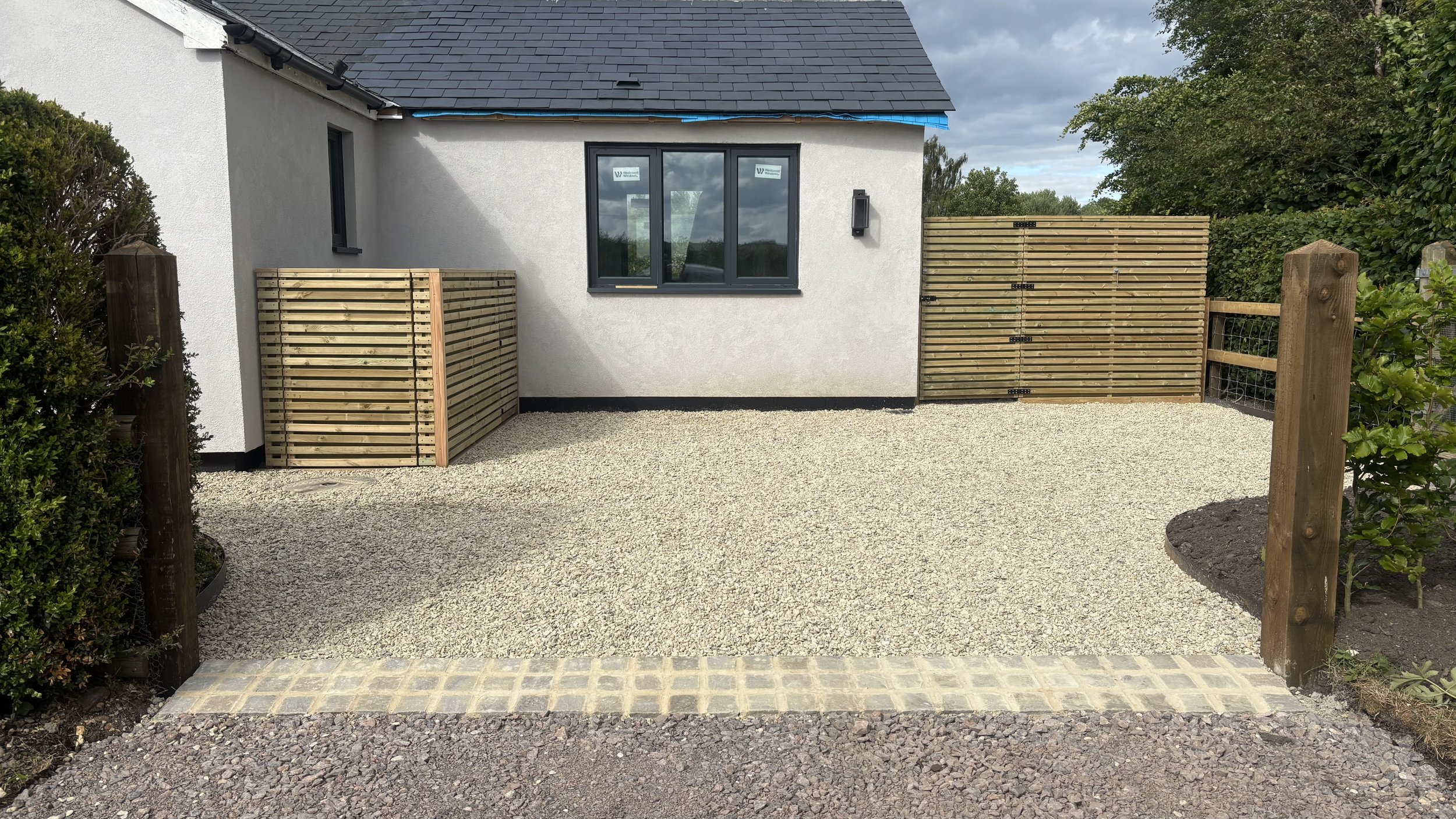 View of a gravel driveway with a white house, bespoke Venetian style bin store screening, sandstone setts edging driveway steel flower bed edging and some green shrubbery and trees.