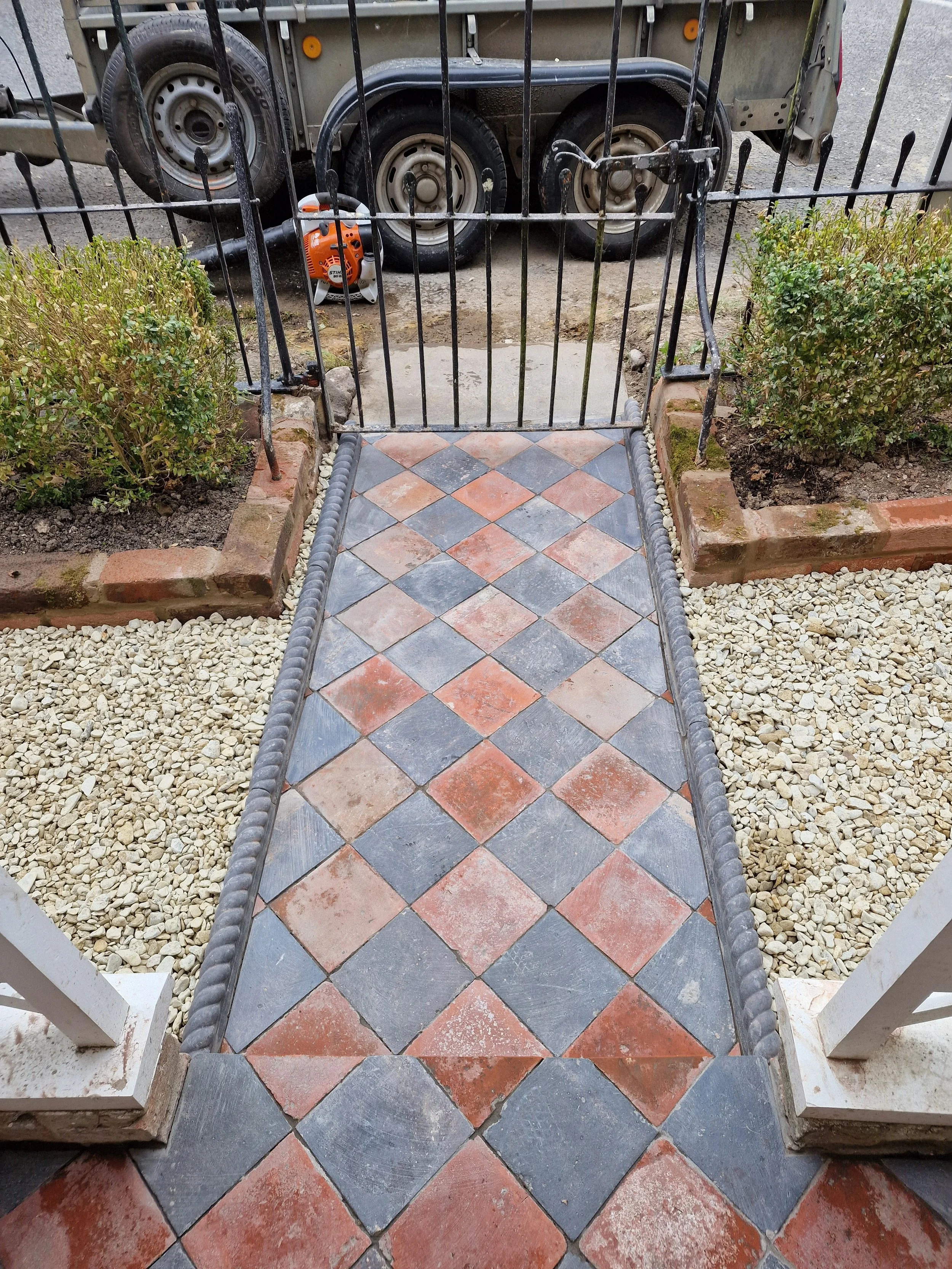 A small set of tile stairs leading to a porch with gardan plants on either side. A black metal gate is partly open.