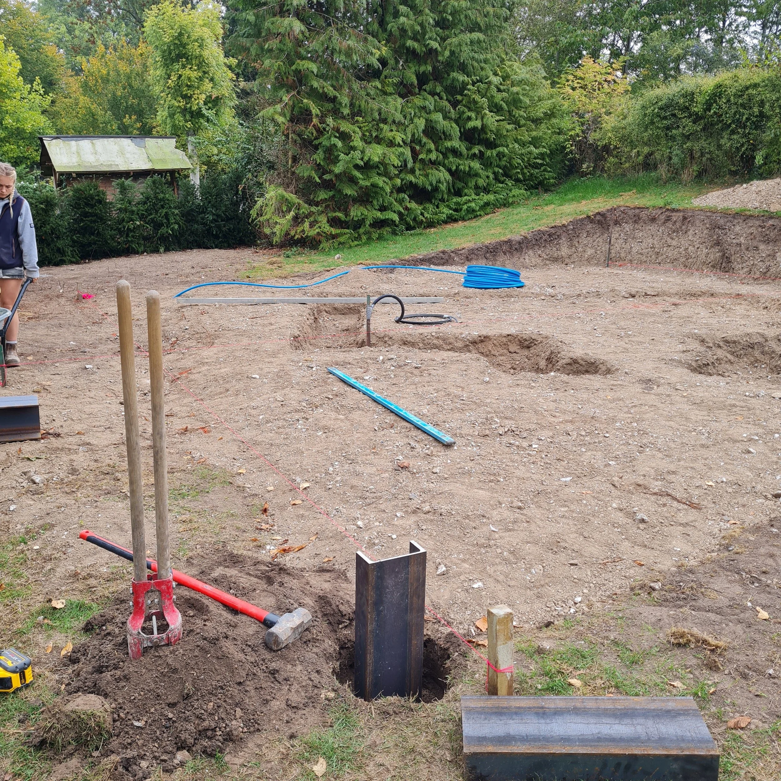 A construction site outdoors with an excavated hole, construction tools, and pipes, surrounded by green trees and grass.
