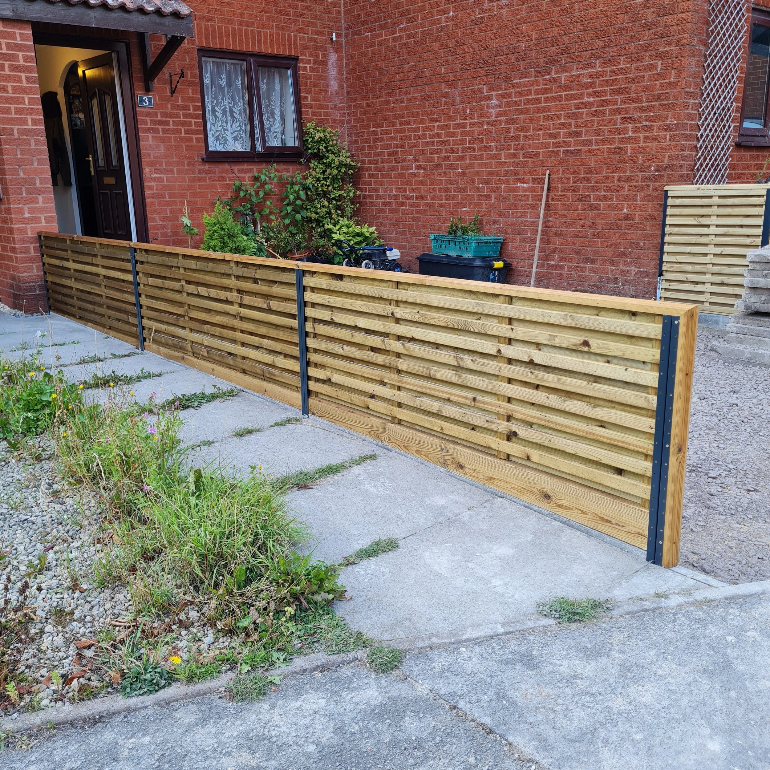 A small urban garden with plants in pots next to a brick wall, a black compost bin, and a newly installed wooden fence.