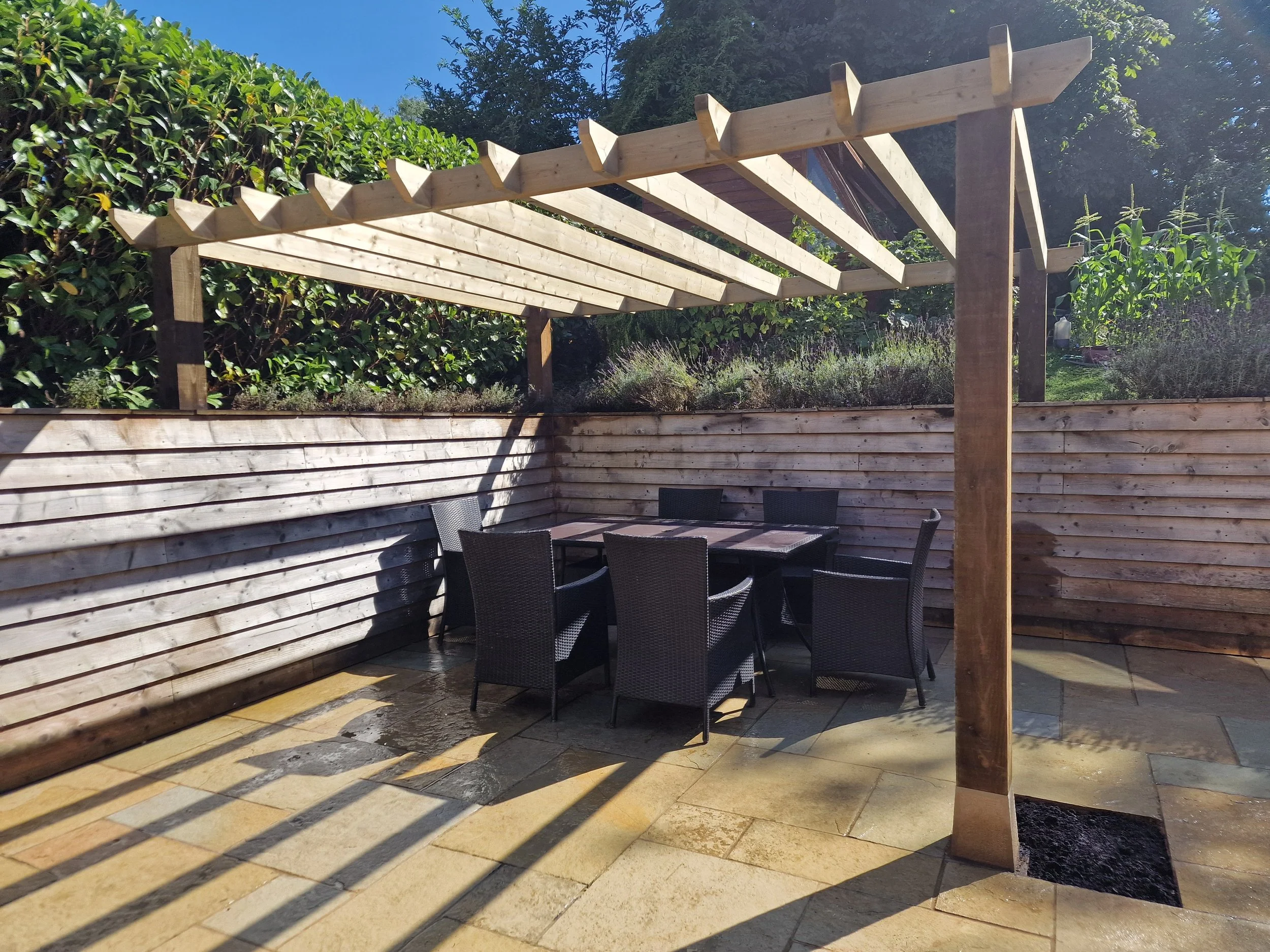 Outdoor patio with black table and chairs, wooden pergola frame, wooden fence, potted plants, lush green hedge, and blue sky.