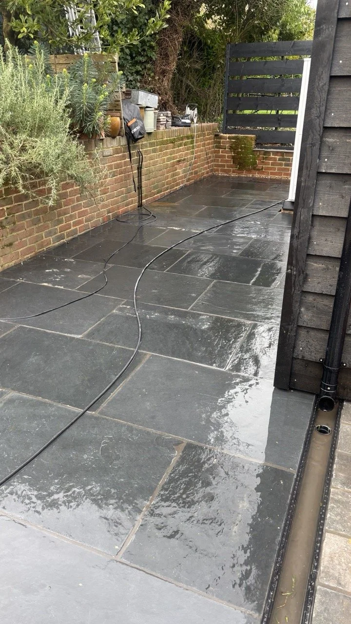 Wet black stone patio with a black garden hose and a black extension cord, bordered by a brick wall with greenery and a black wooden fence.