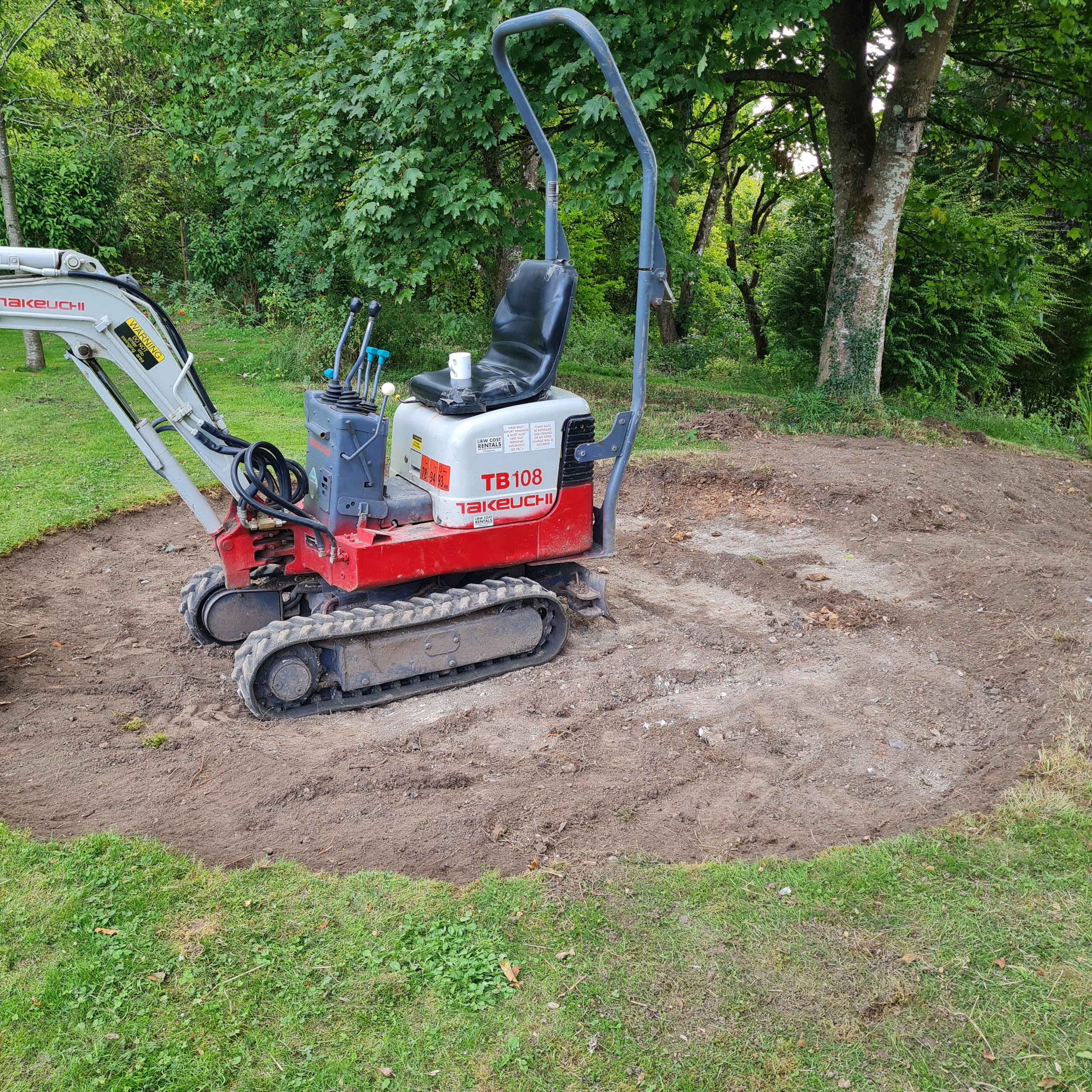 A small white and red compact excavator with a black seat, controls, and tracks, positioned outdoors on dirt ground surrounded by green grass, trees, and foliage.
