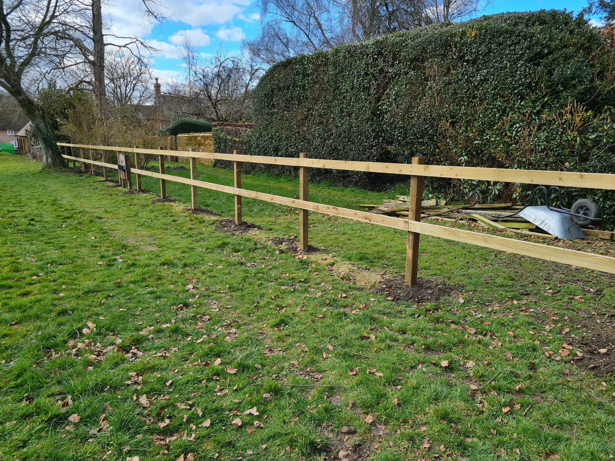A grassy backyard with a newly constructed wooden fence, a large hedge, trees, and a small building in the background under a partly cloudy sky.