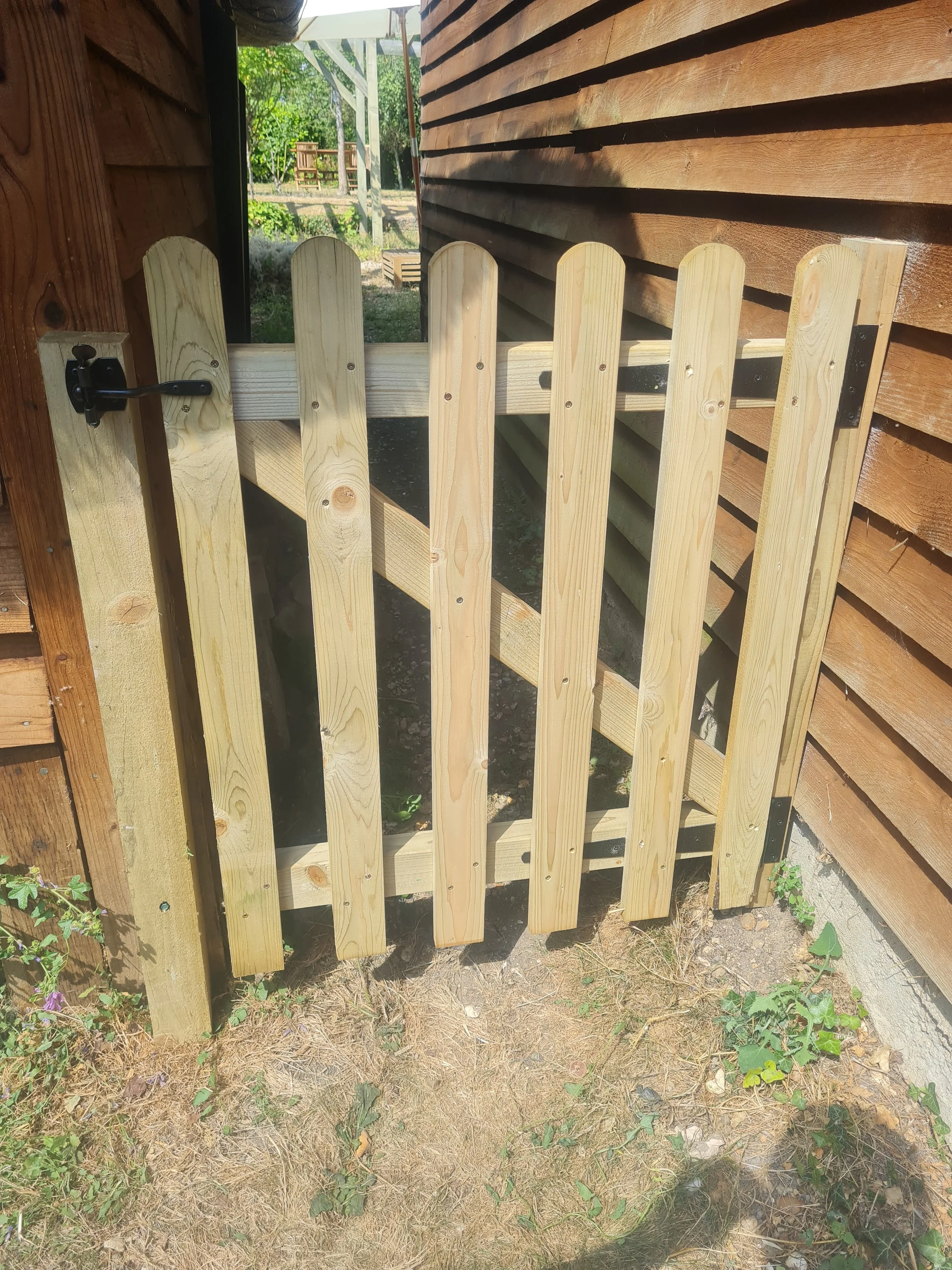 A wooden fence with a latch is in front of a wooden wall. A garden with trees and a bench are visible in the background.