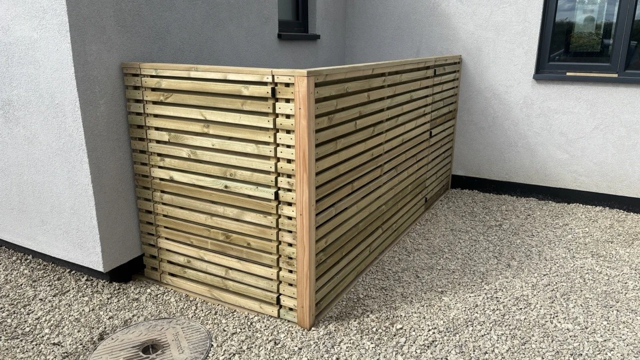 A newly built wooden privacy fence attached to the corner of a house, located on a gravel yard with a round manhole cover nearby.