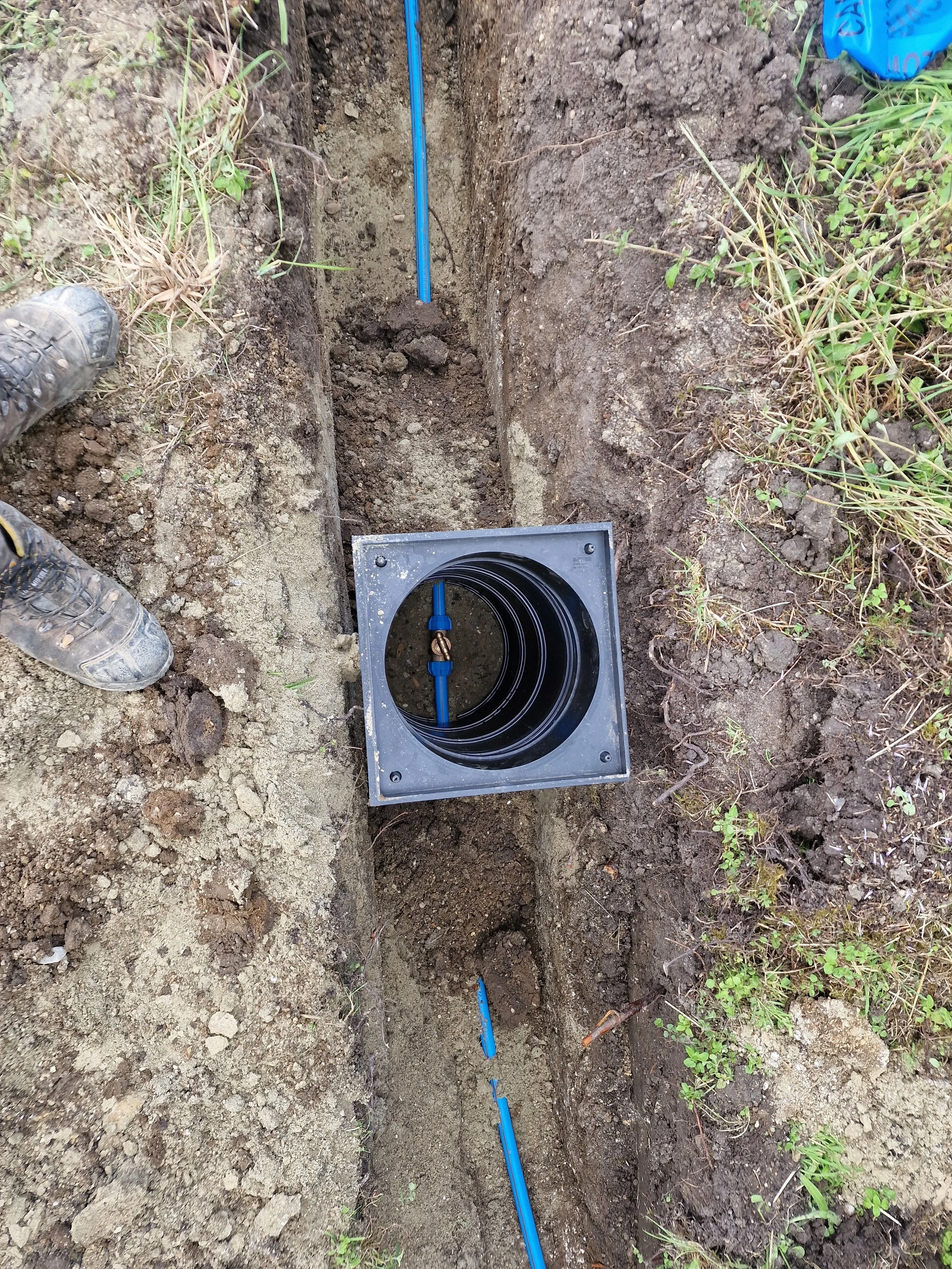 A trench dug in the ground with blue piping and a black plastic conduit installed, surrounded by dirt and grass.